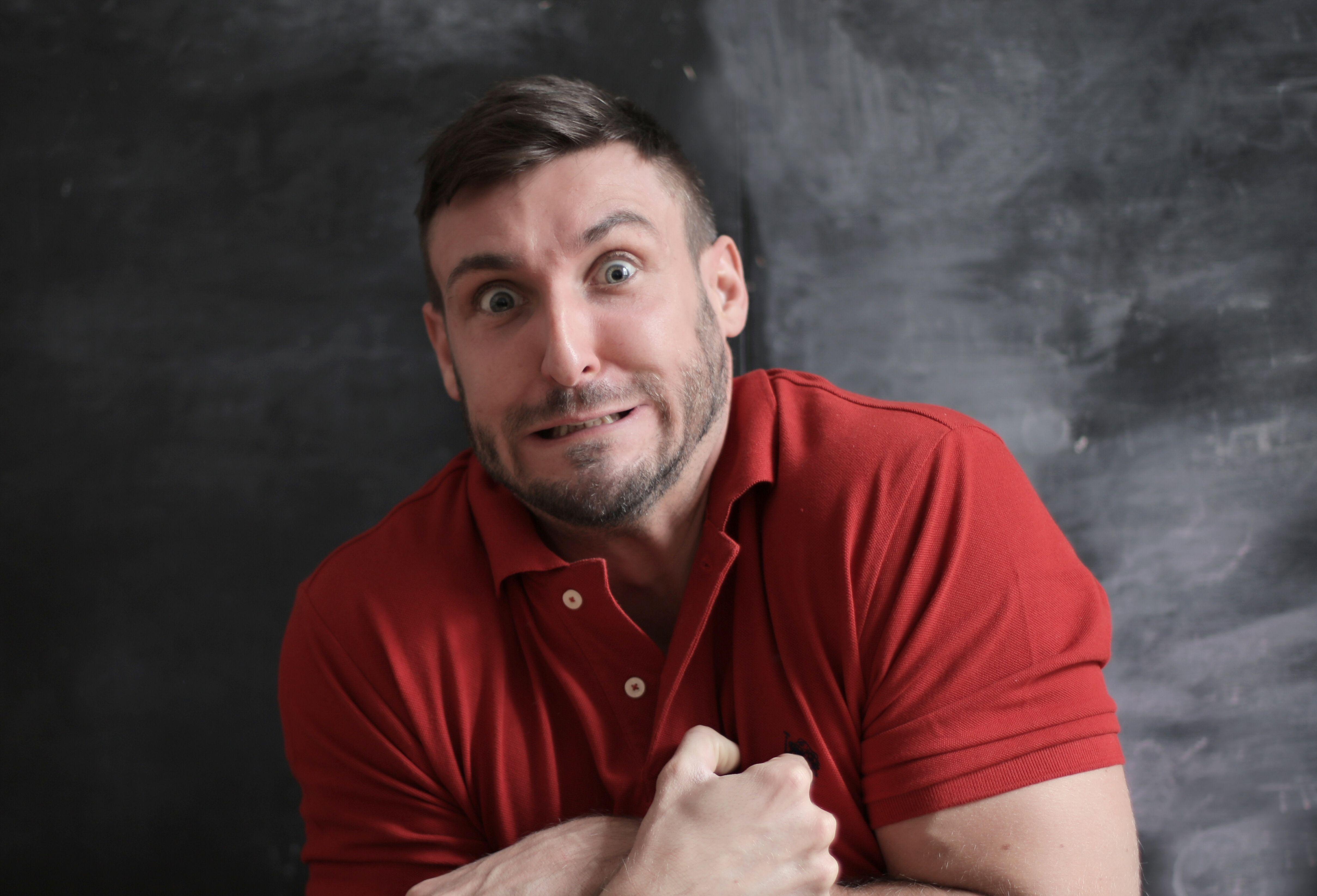 Man in red shirt making a humorous, exaggerated facial expression against a dark background.