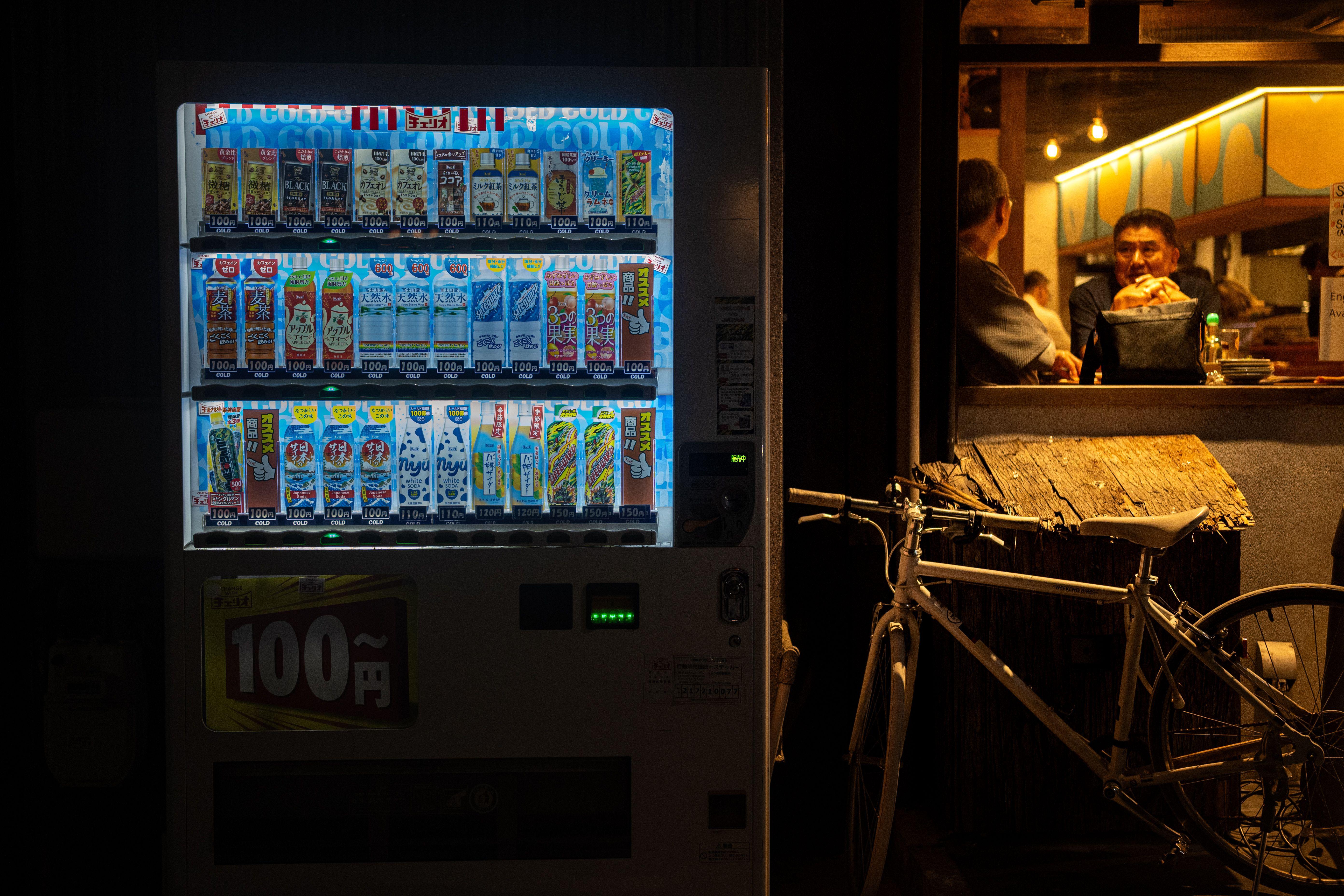 Vending machine glowing at night beside a cozy café with two men inside.