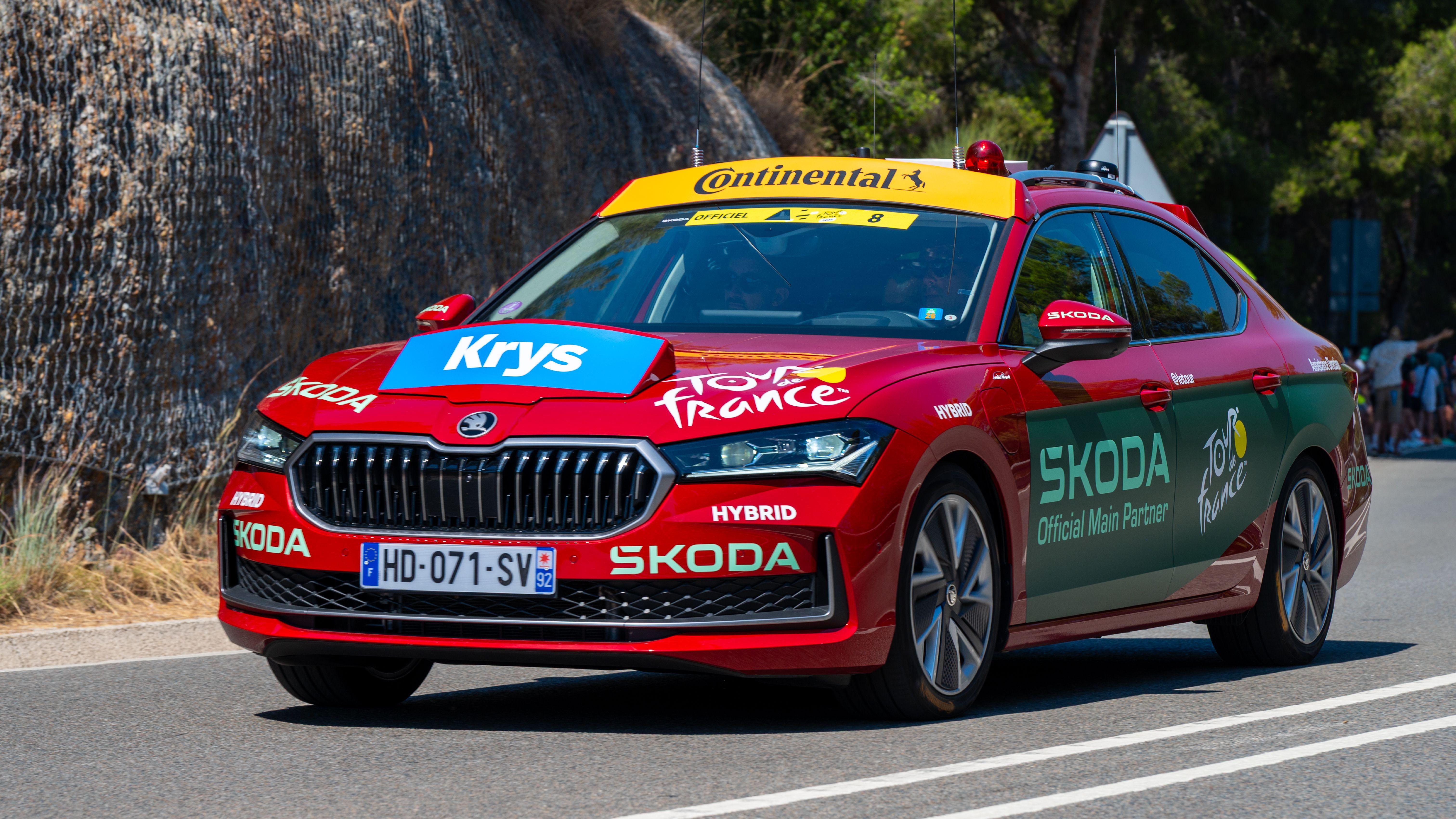 Red ŠKODA support car at Tour de France with branded decals on scenic route.
