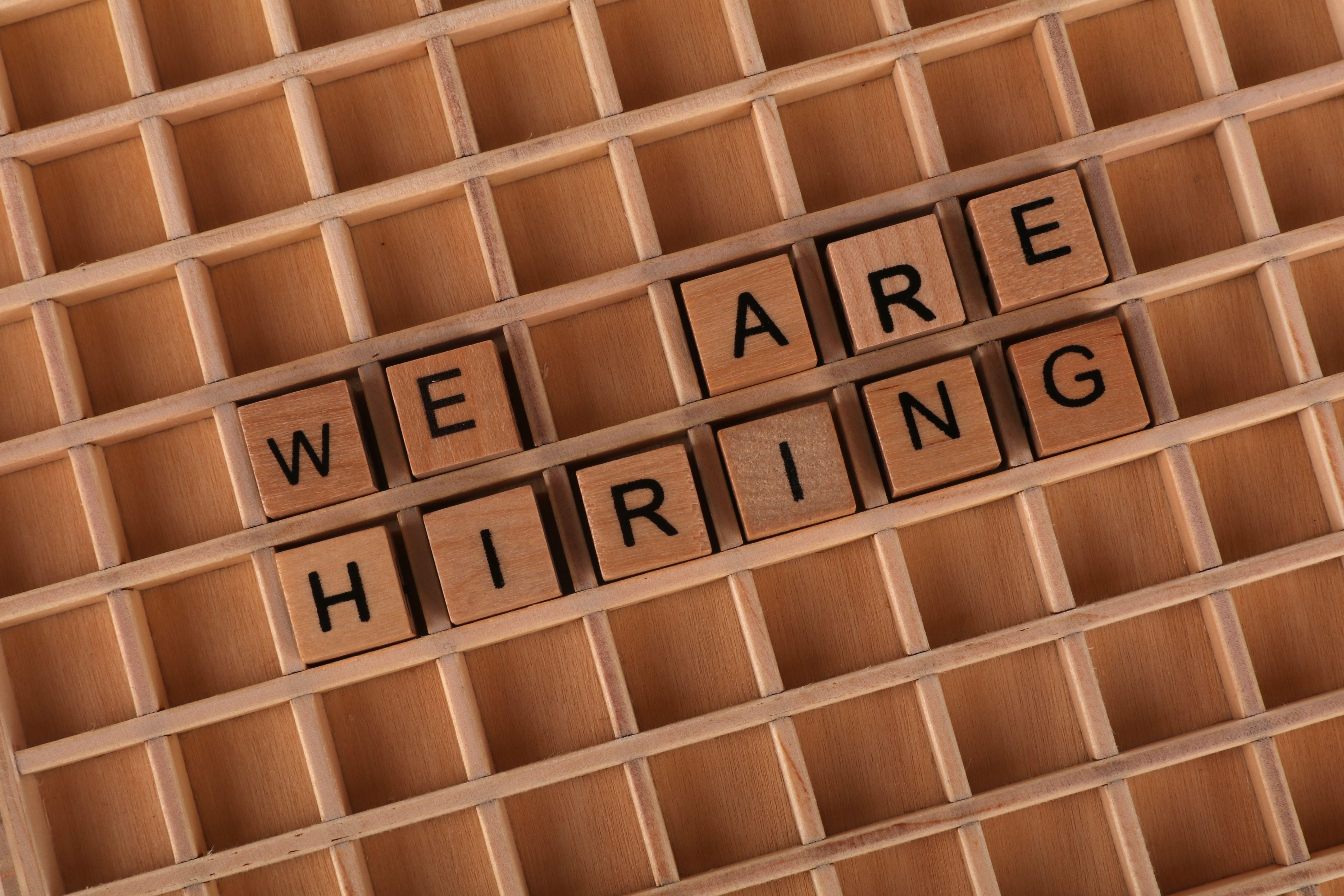 Wooden letter tiles arranged to spell 'We Are Hiring' on a wooden grid background.