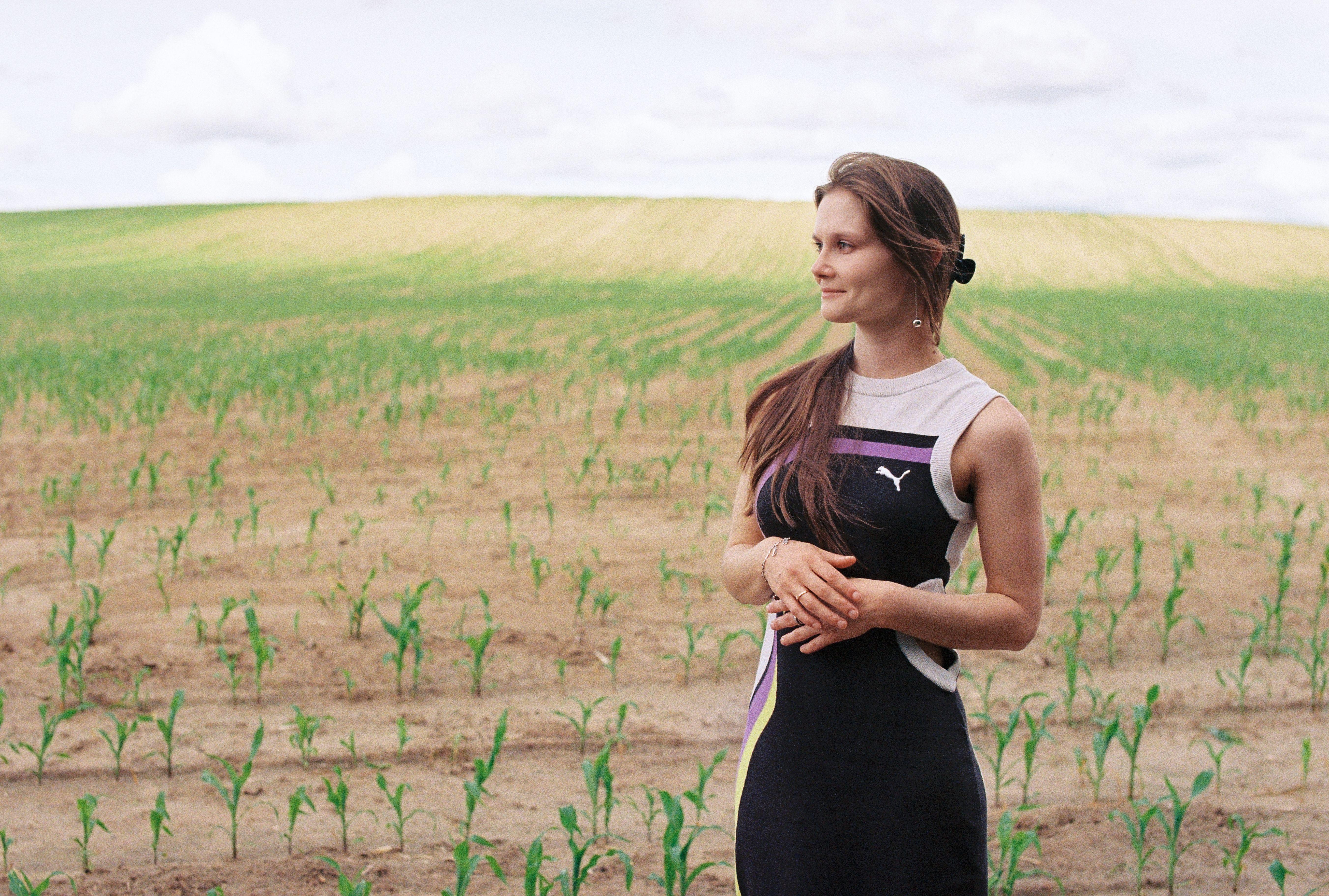A young woman stands thoughtfully in a vast field under a bright summer sky.