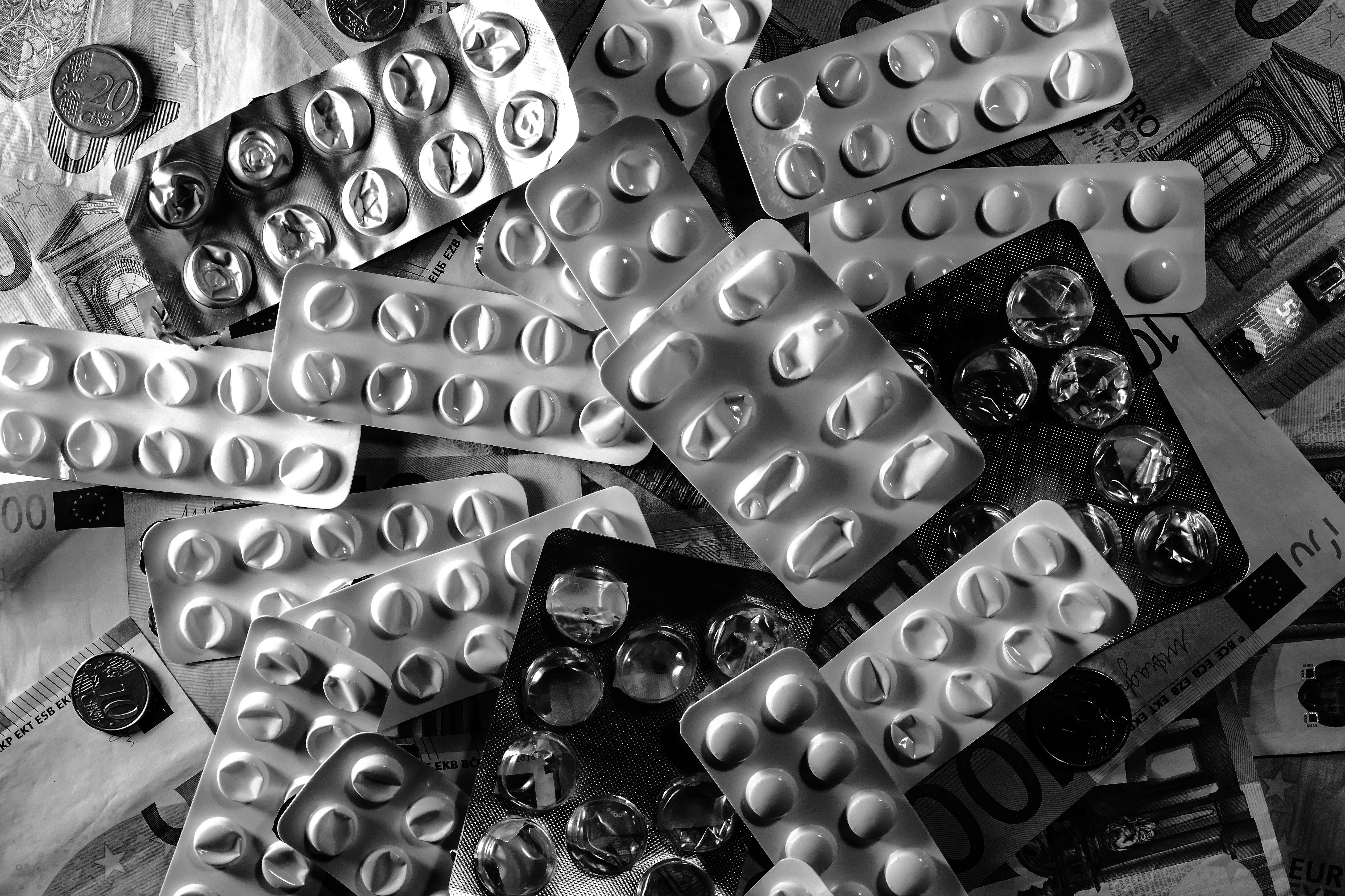 Black and white image of pill blister packs scattered on euro bills and coins.