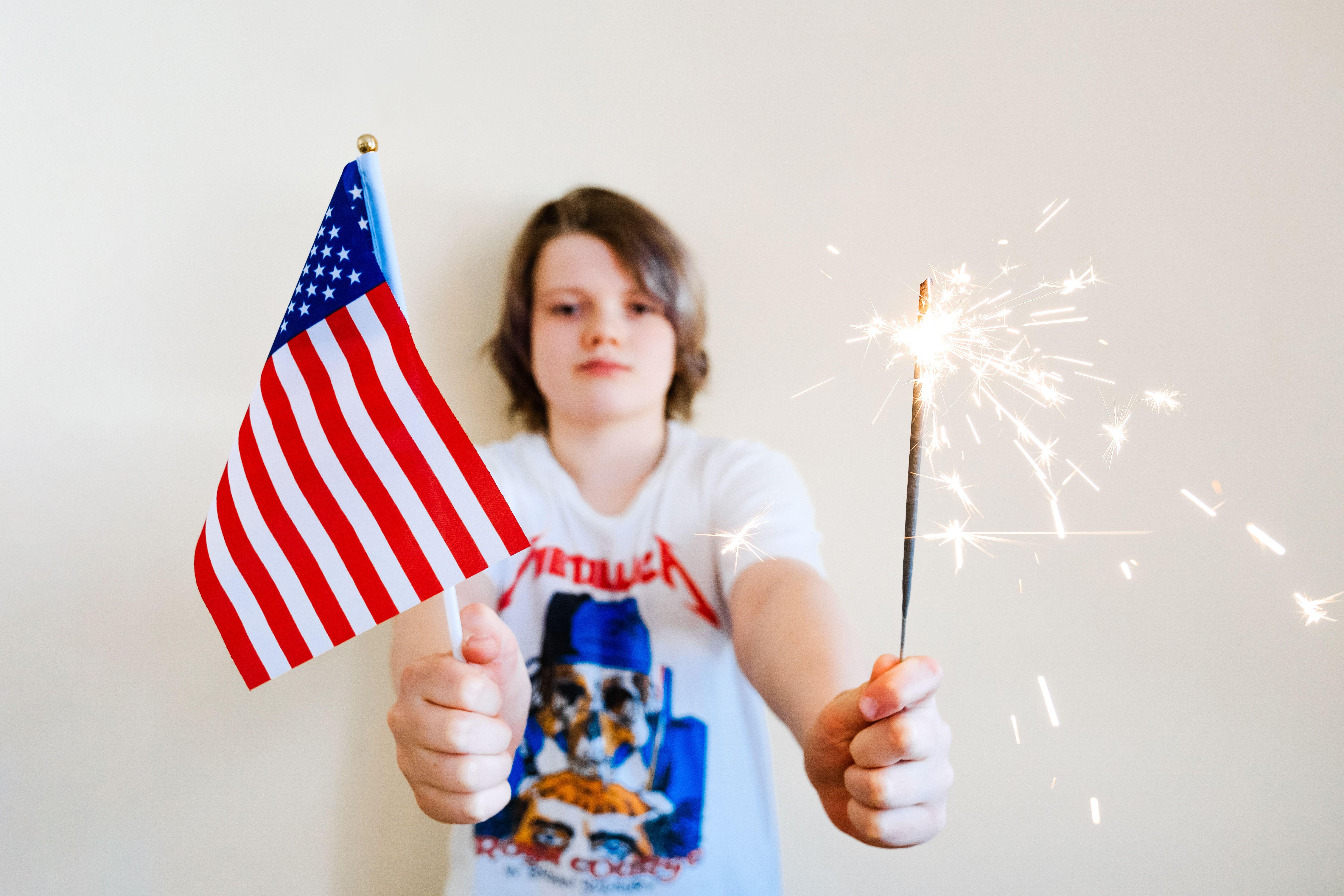 Young child joyfully holding American flag and sparkler indoors, celebrating USA holidays.
