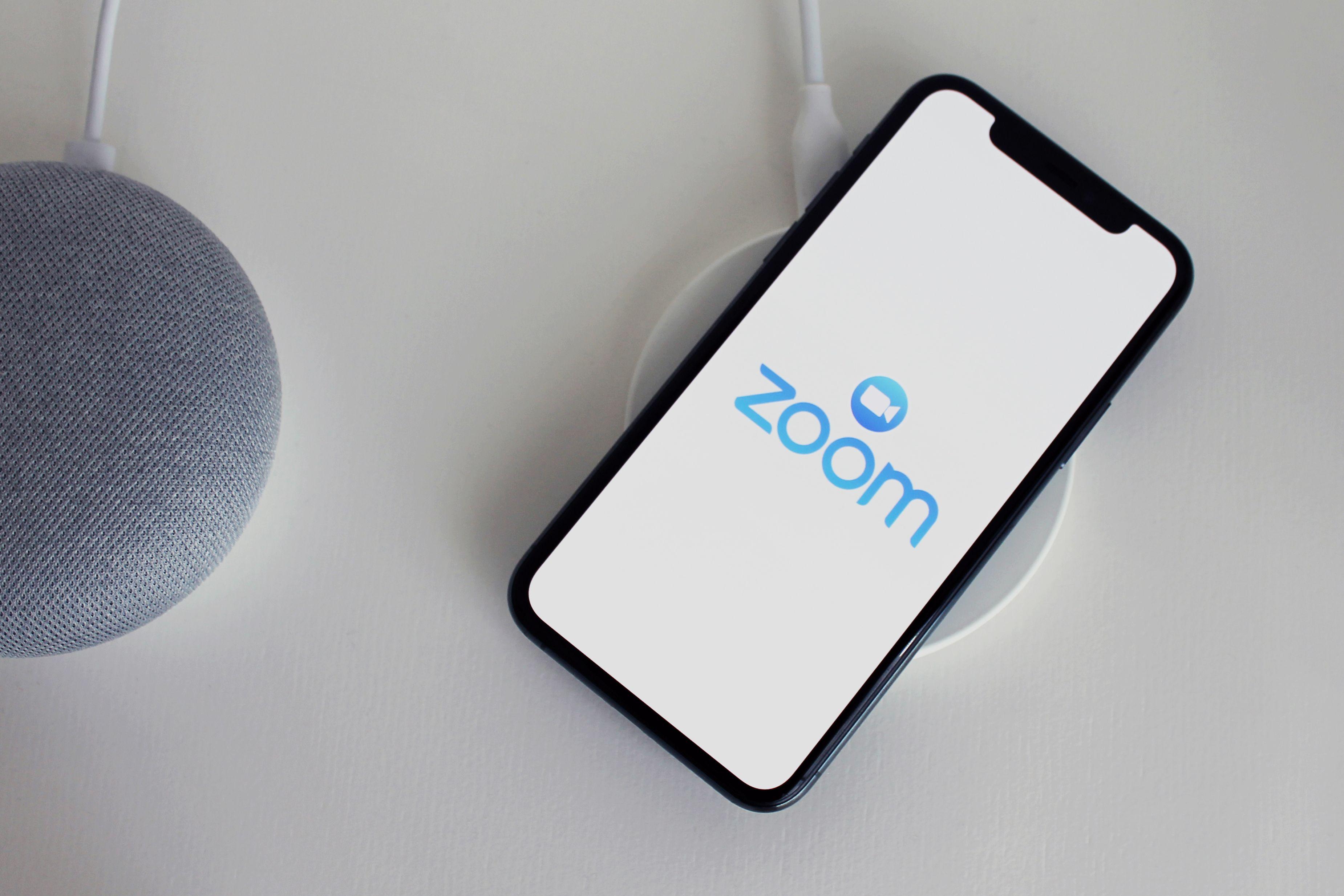 Close-up of a smartphone with Zoom app on a wireless charger next to a smart speaker.
