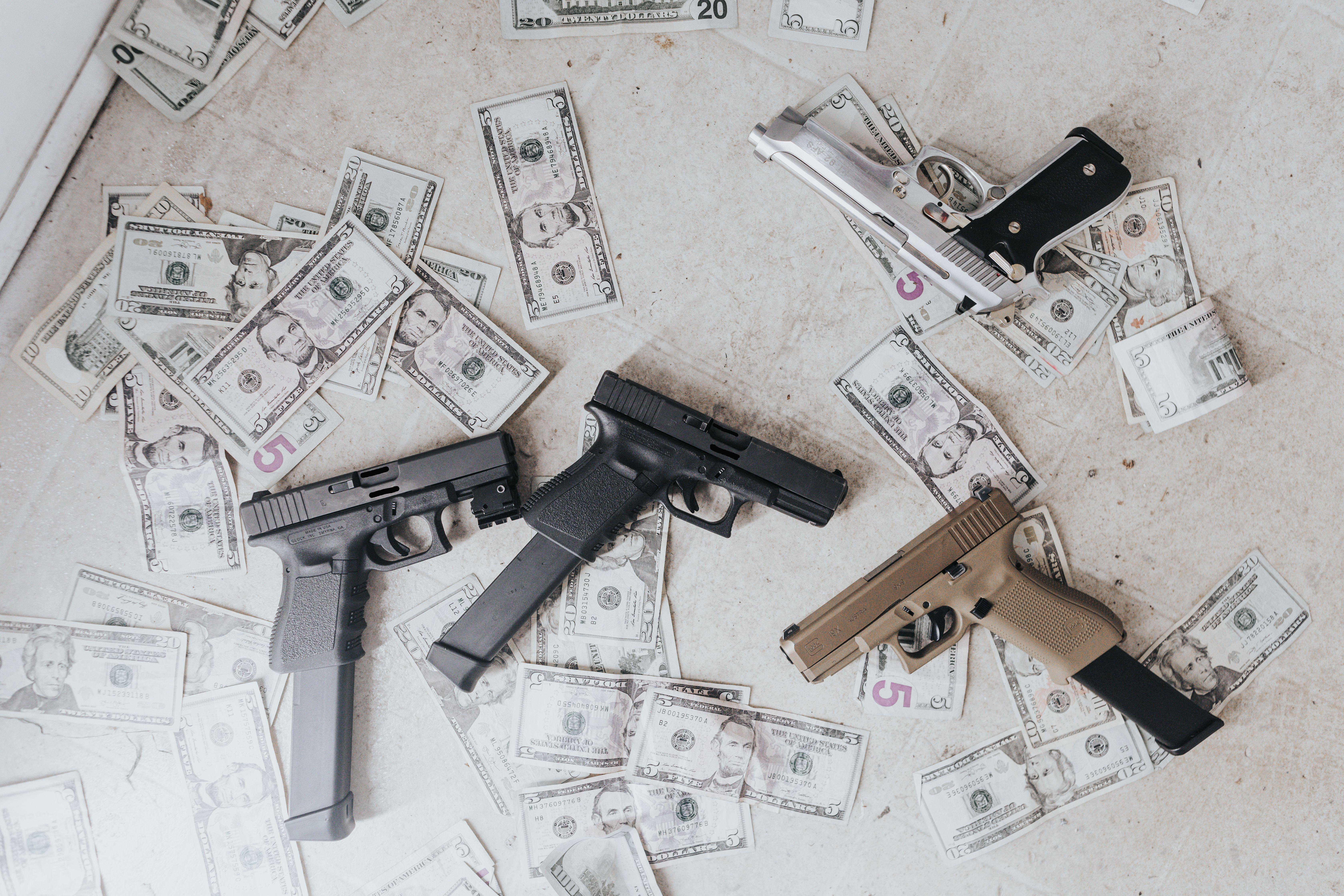 Multiple firearms and US dollar bills scattered on a floor in a high-angle view.