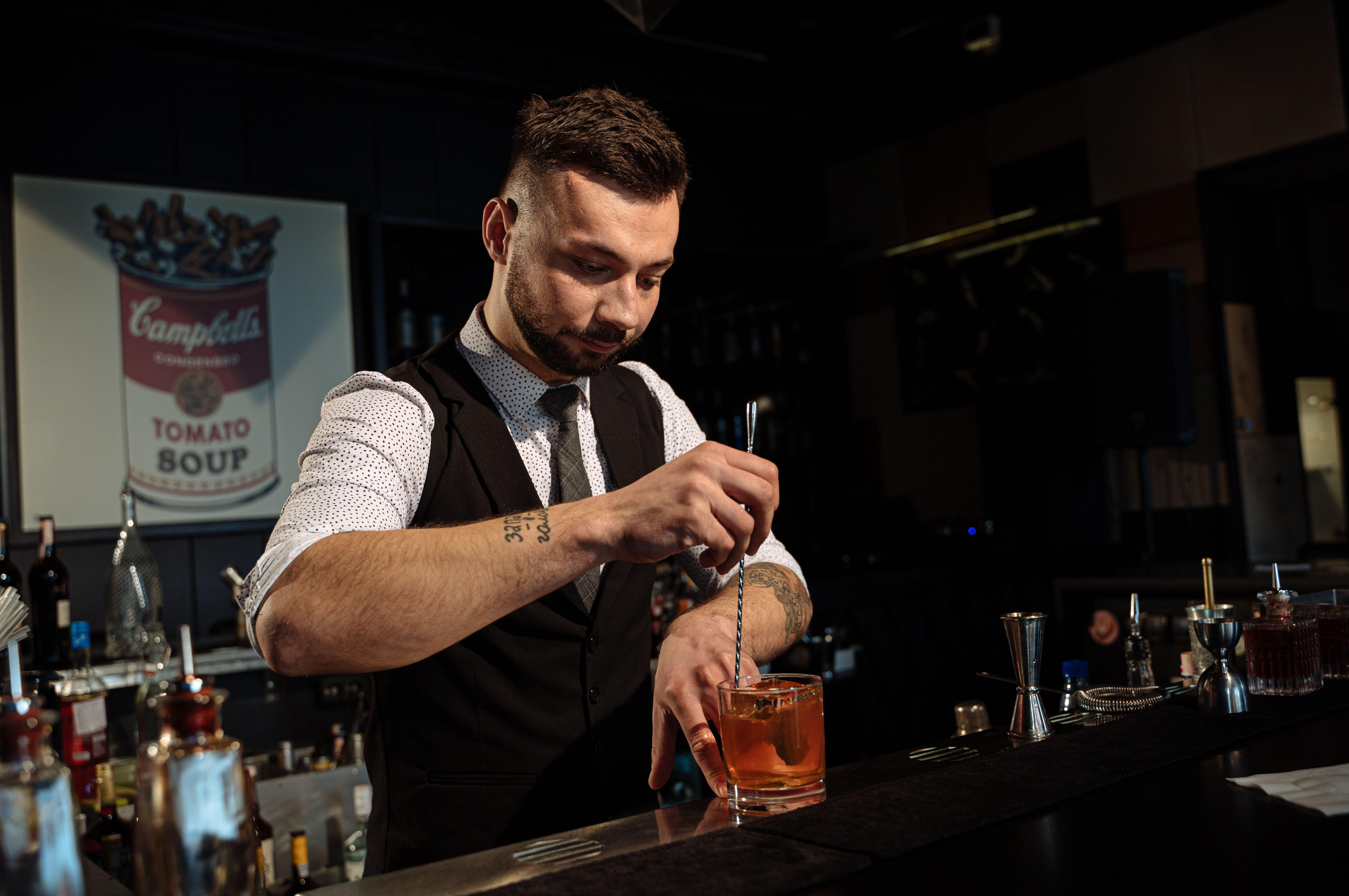 Bartender skillfully mixing a cocktail at a stylish bar with a Campbell's Soup poster in the background.