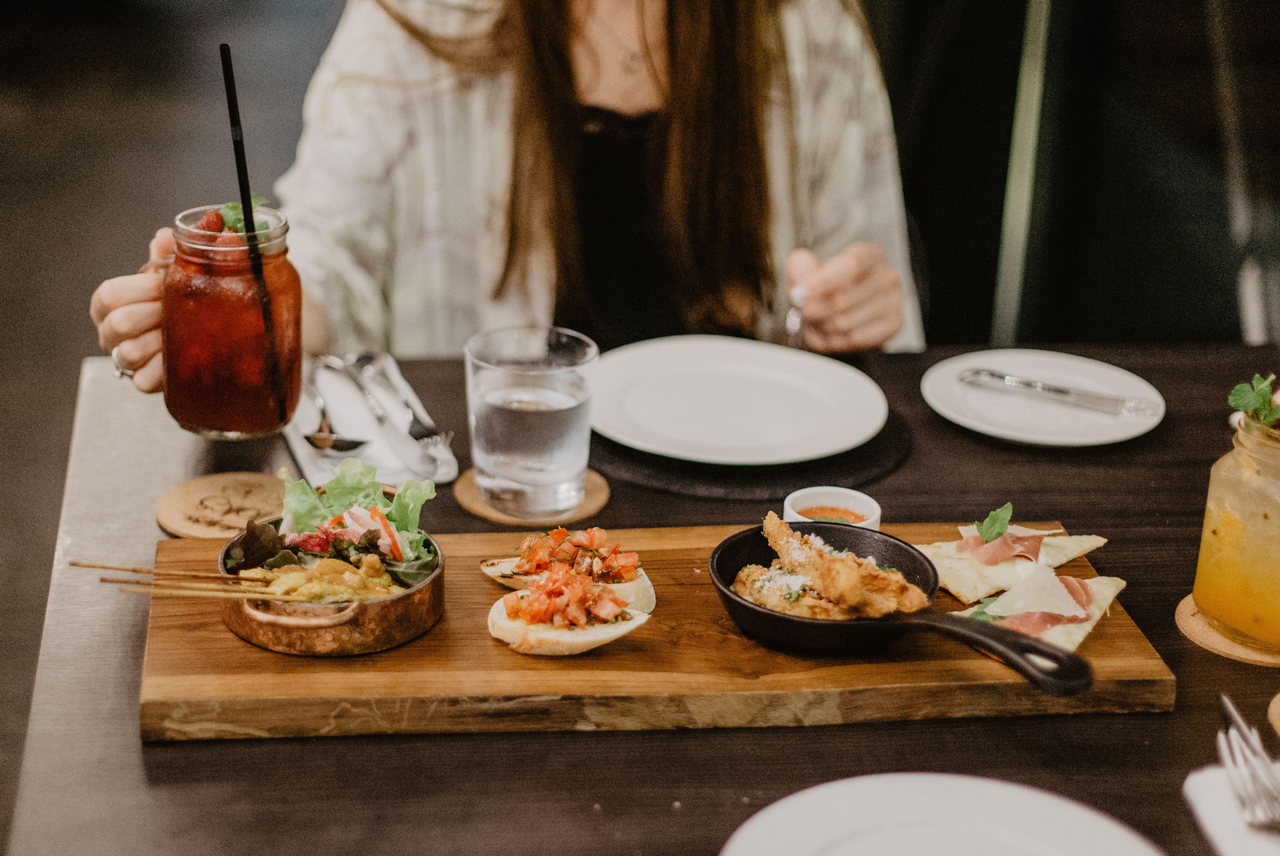Crop unrecognizable female in casual clothes having dinner in Asian restaurant while sitting at table with berry mocktail and grilled meat on sticks served with tomato sandwiches and spring rolls