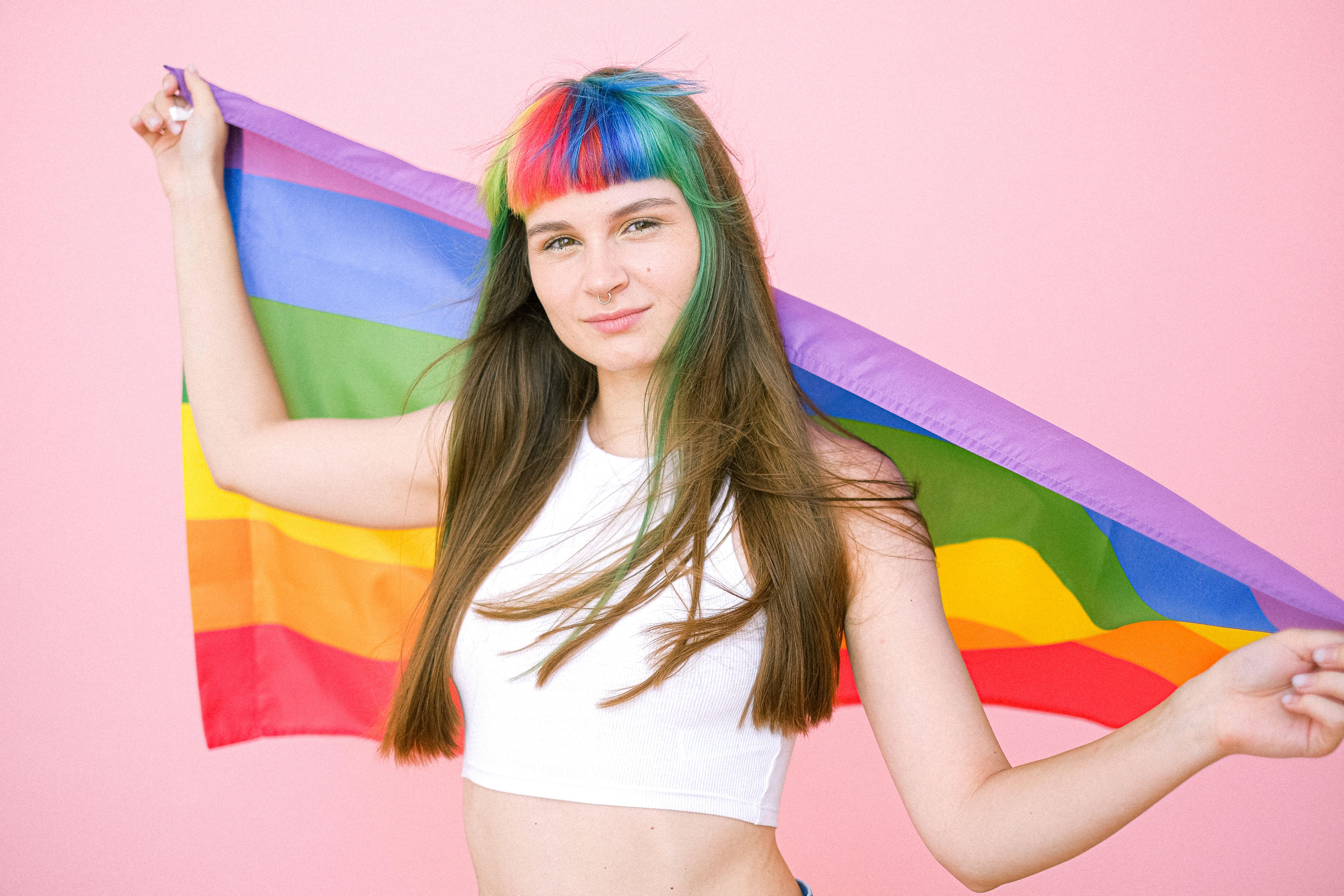 A woman with rainbow bangs holding a pride flag against a pink background, symbolizing LGBTQ pride.