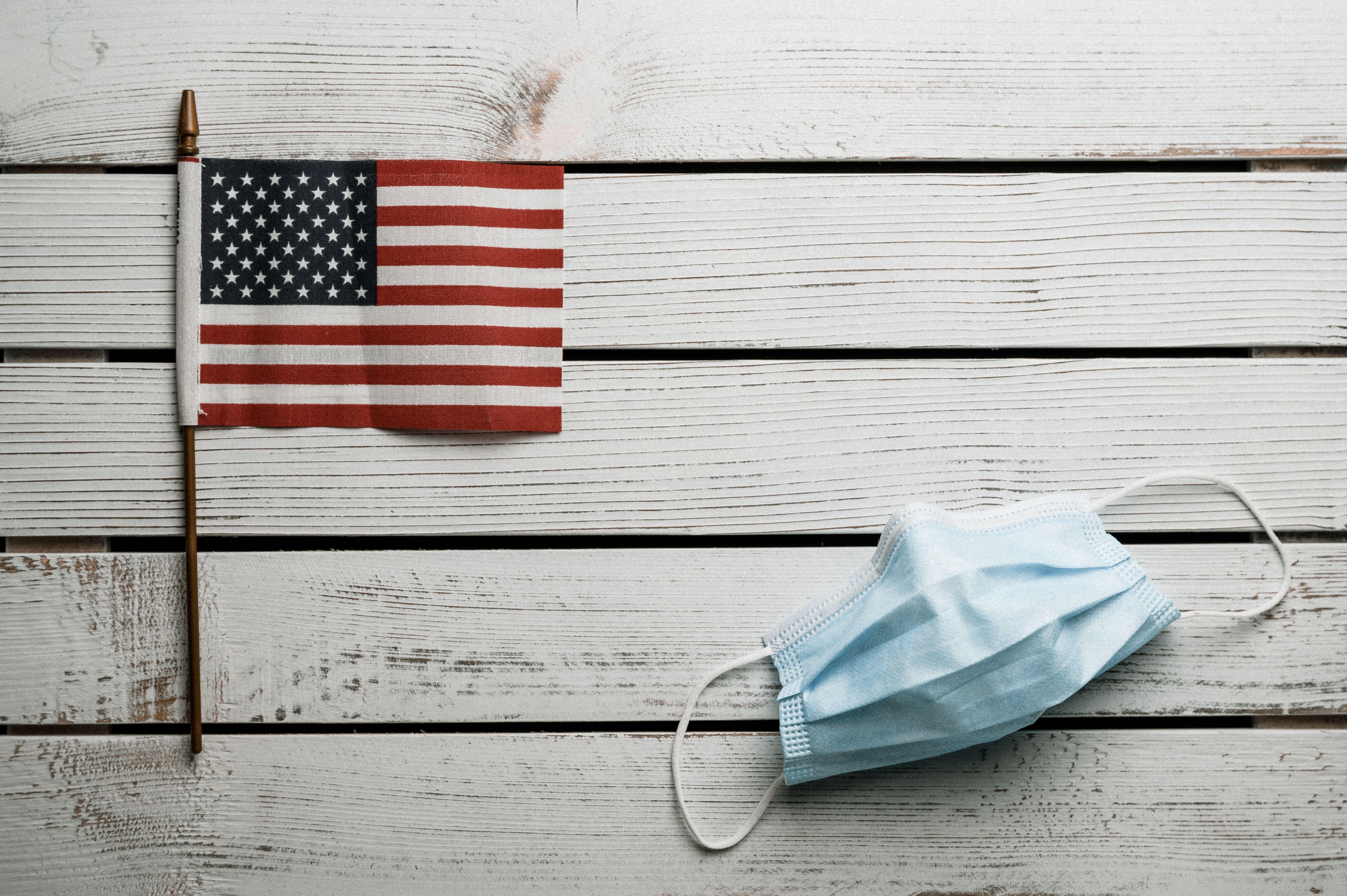 Top view of small Flag of United States and light blue fabric disposable face mask on white wooden wall