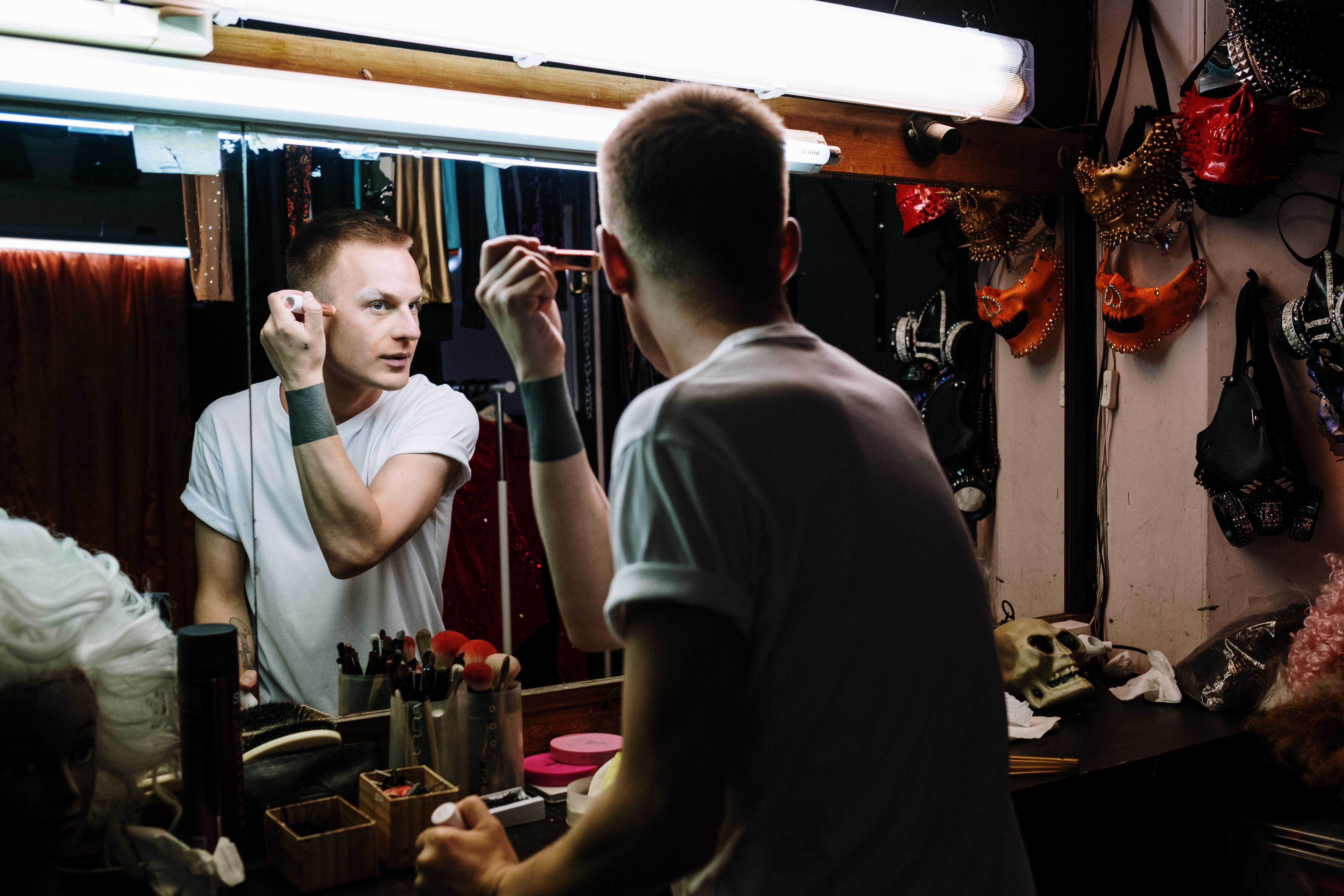 Drag performer applies makeup in a theater dressing room, reflecting transformation and identity.