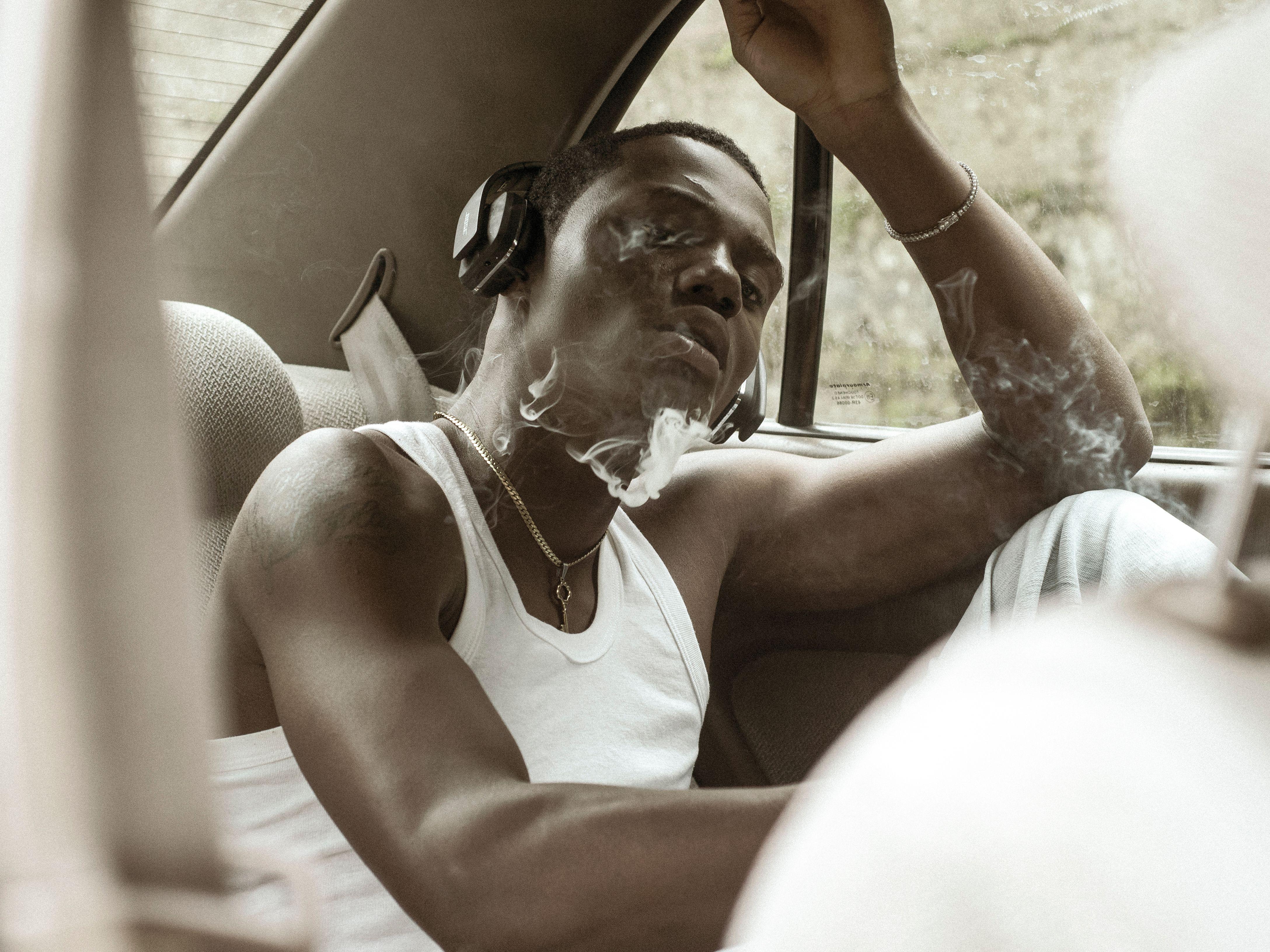 A young man in a car, wearing headphones and inhaling smoke, exudes a calm, introspective mood.