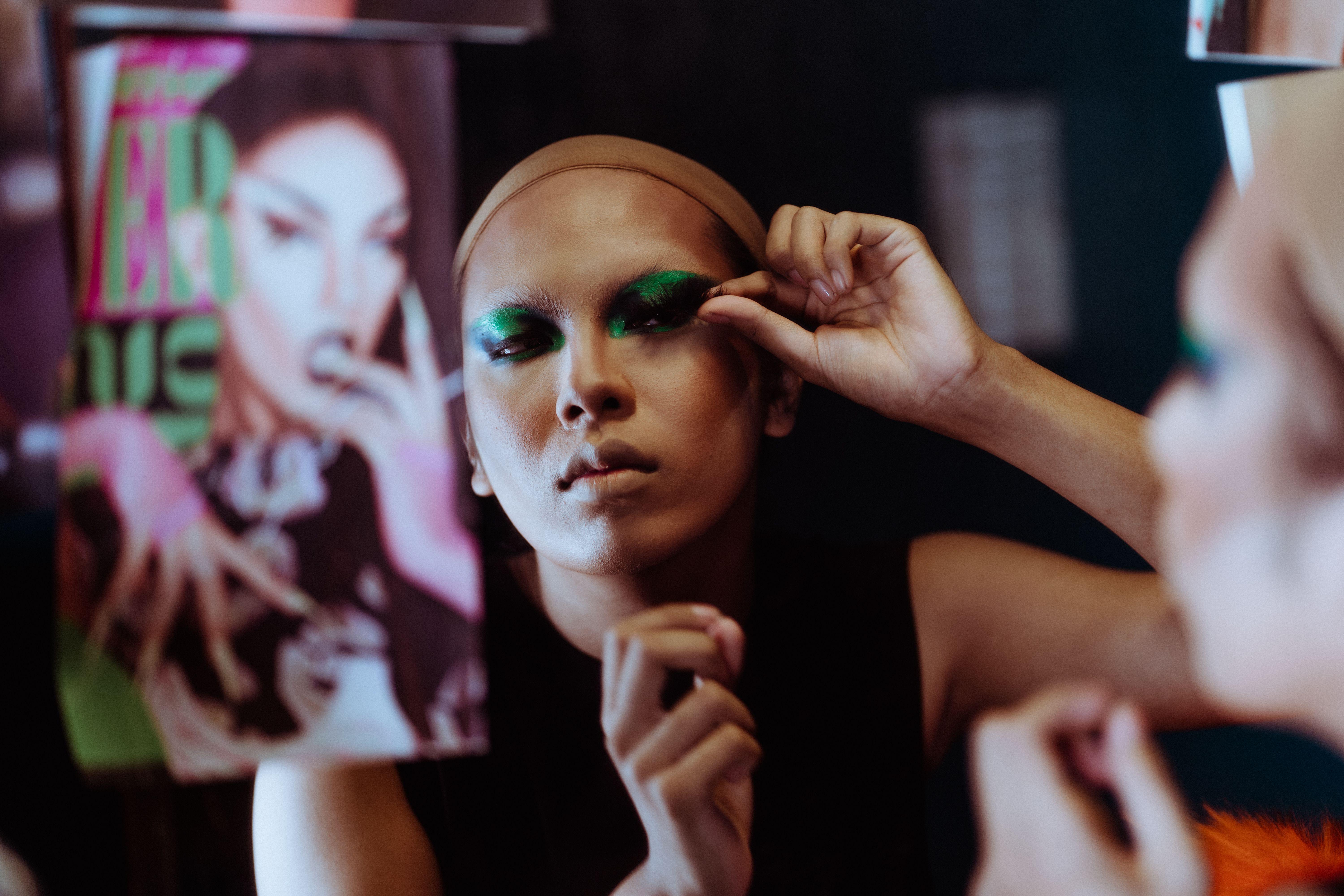 Ethnic female artist in wig cap doing bright makeup with false lashes while looking at mirror reflection