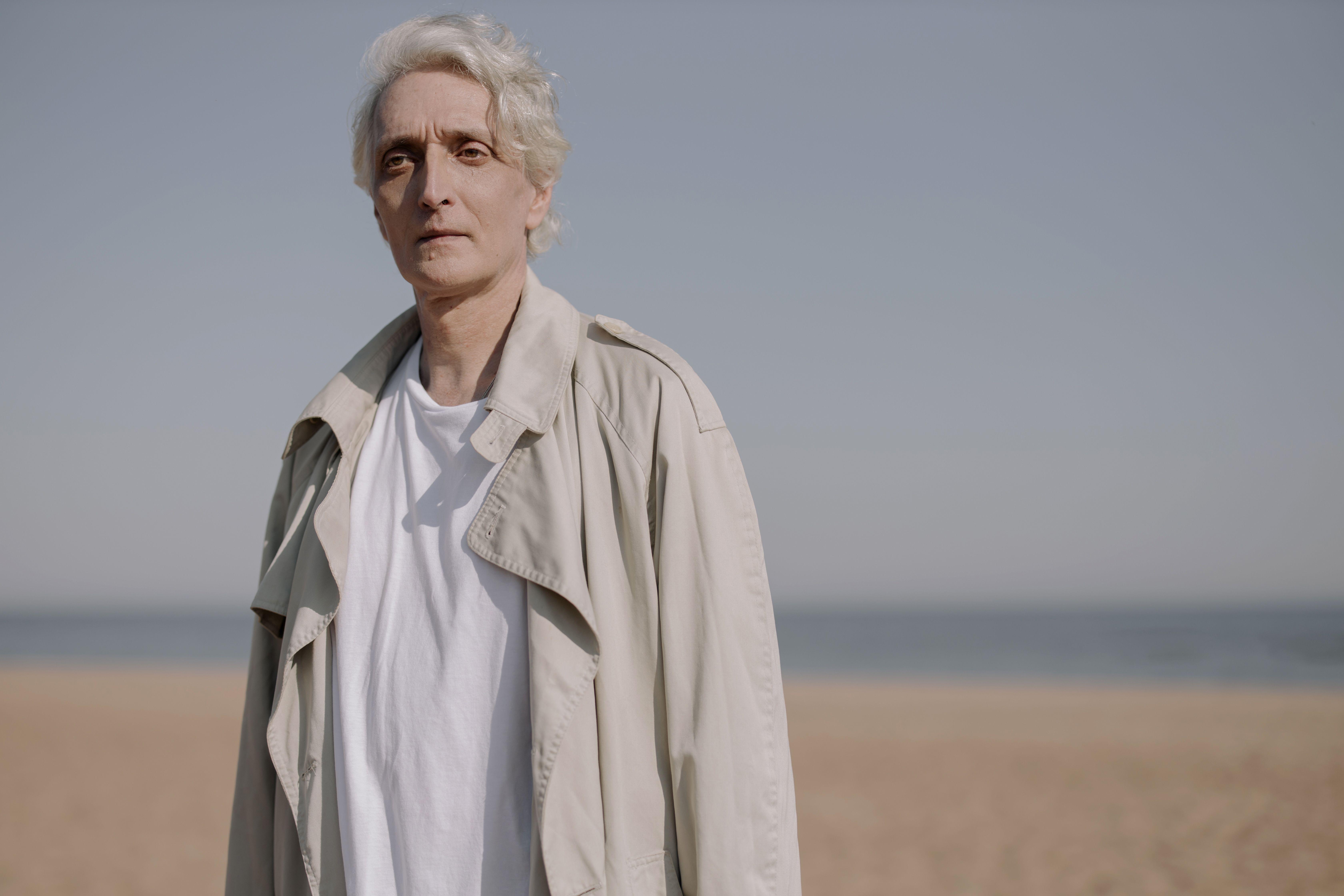 Elderly man in a coat standing on a serene beach under clear skies.