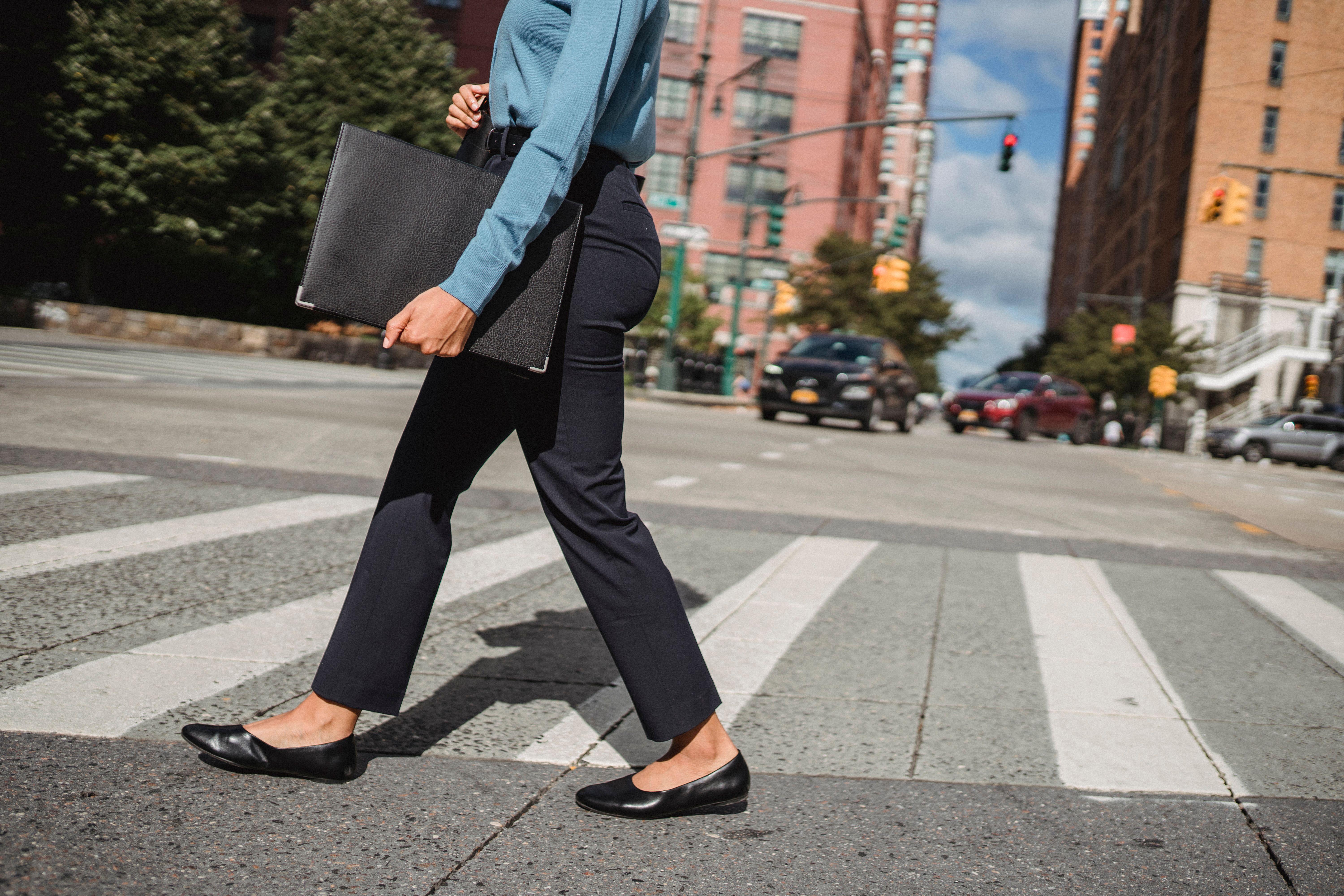 Woman in business attire crossing a street in an urban environment with buildings in the background.