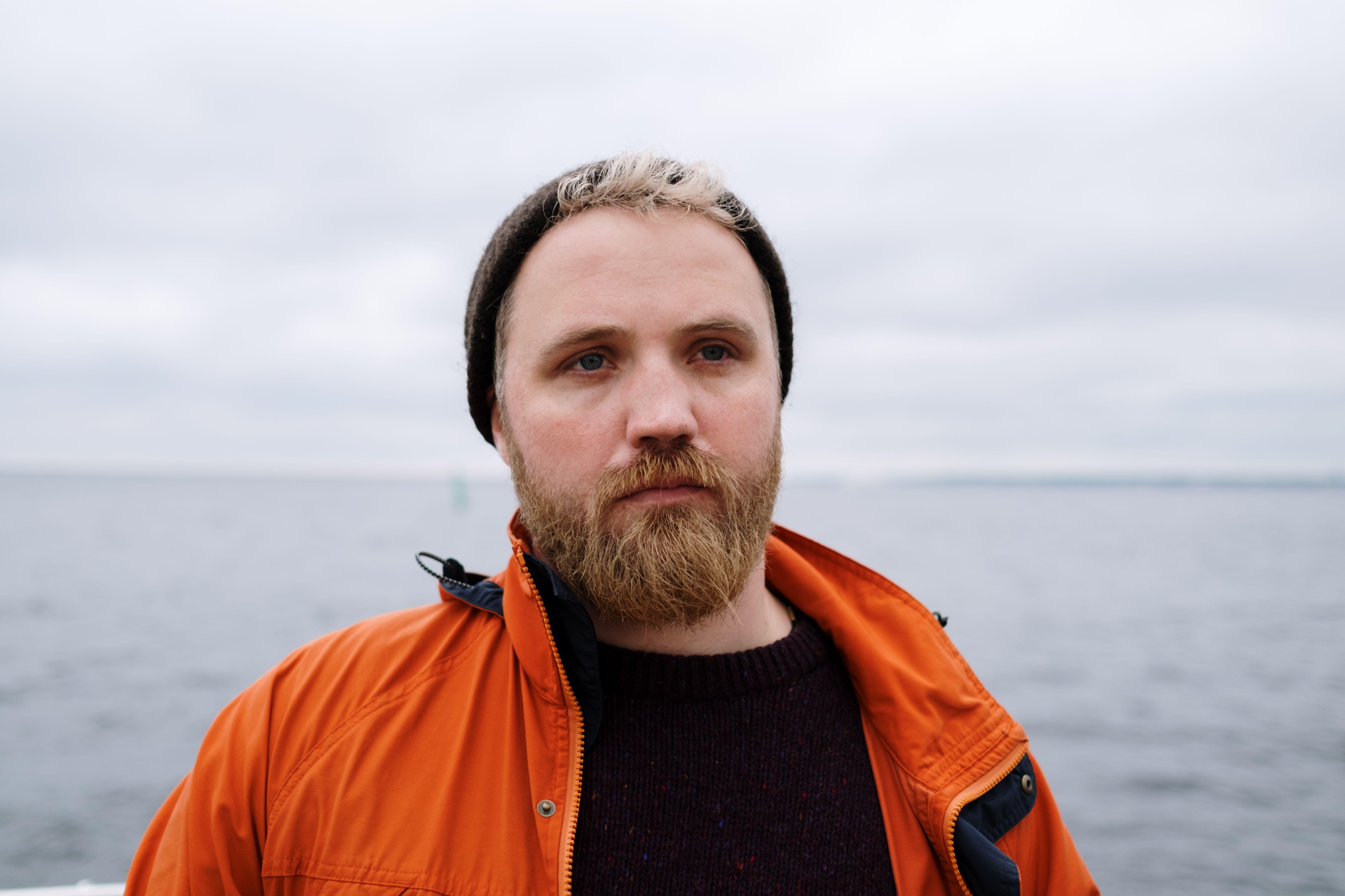 Portrait of a man wearing an orange jacket, standing by the sea with a beard and beanie.