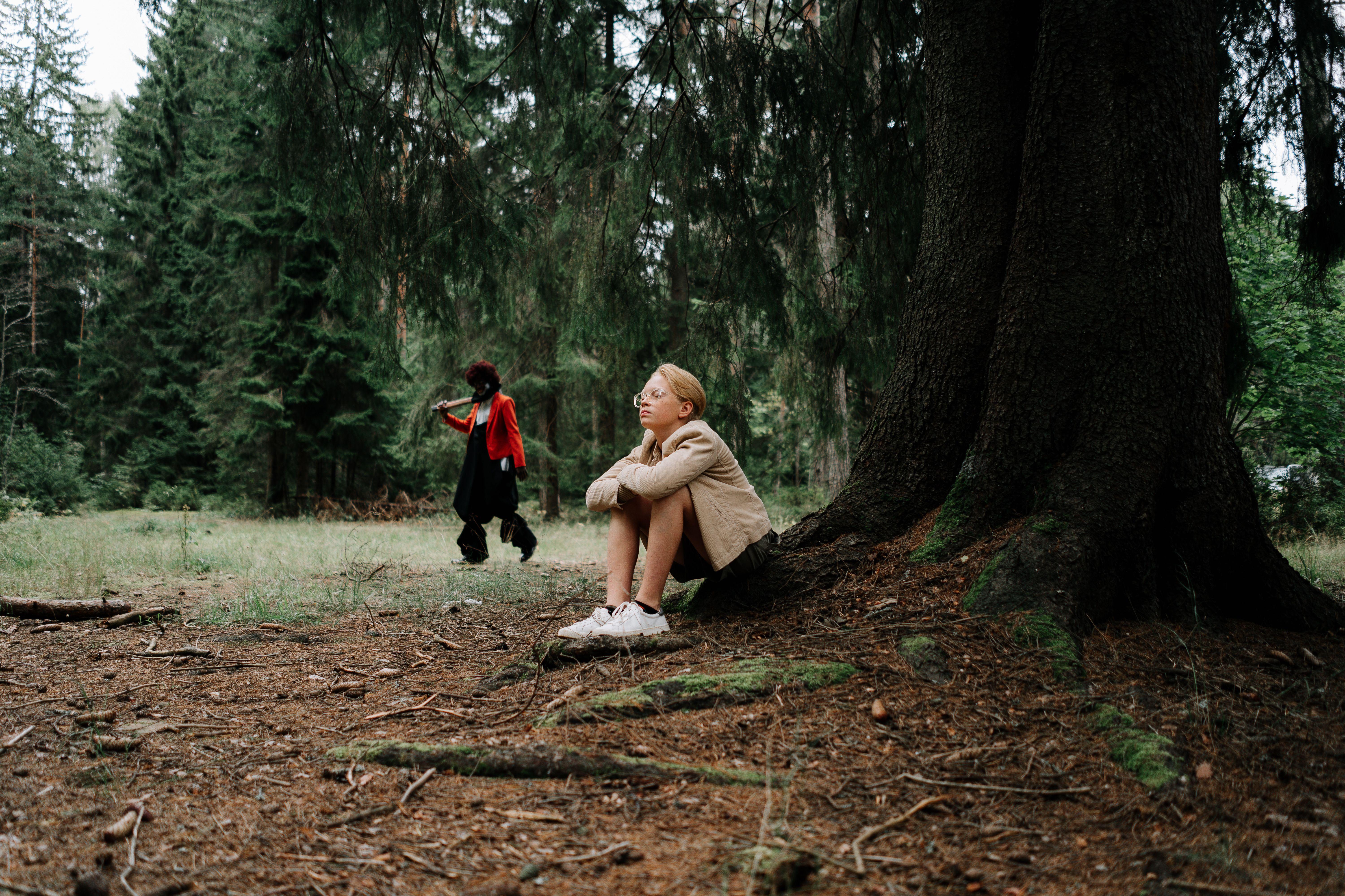 A teenager sits by a tree in a forest, unaware of a masked figure approaching.
