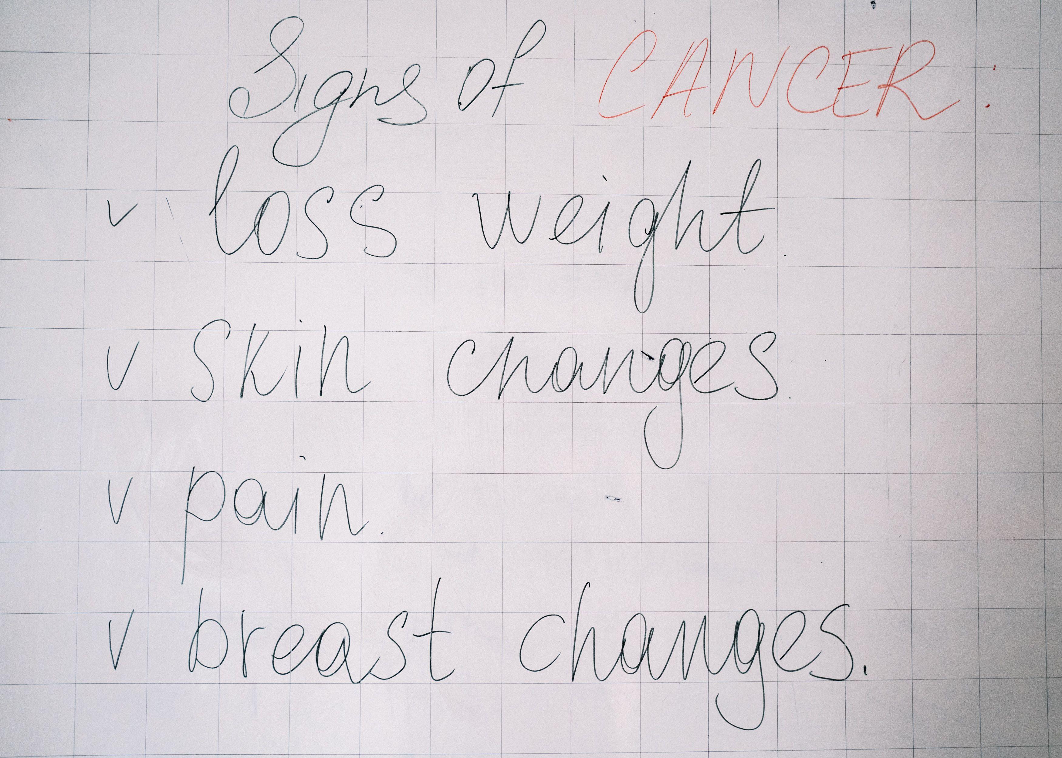 List of cancer symptoms handwritten on a whiteboard including weight loss and skin changes.