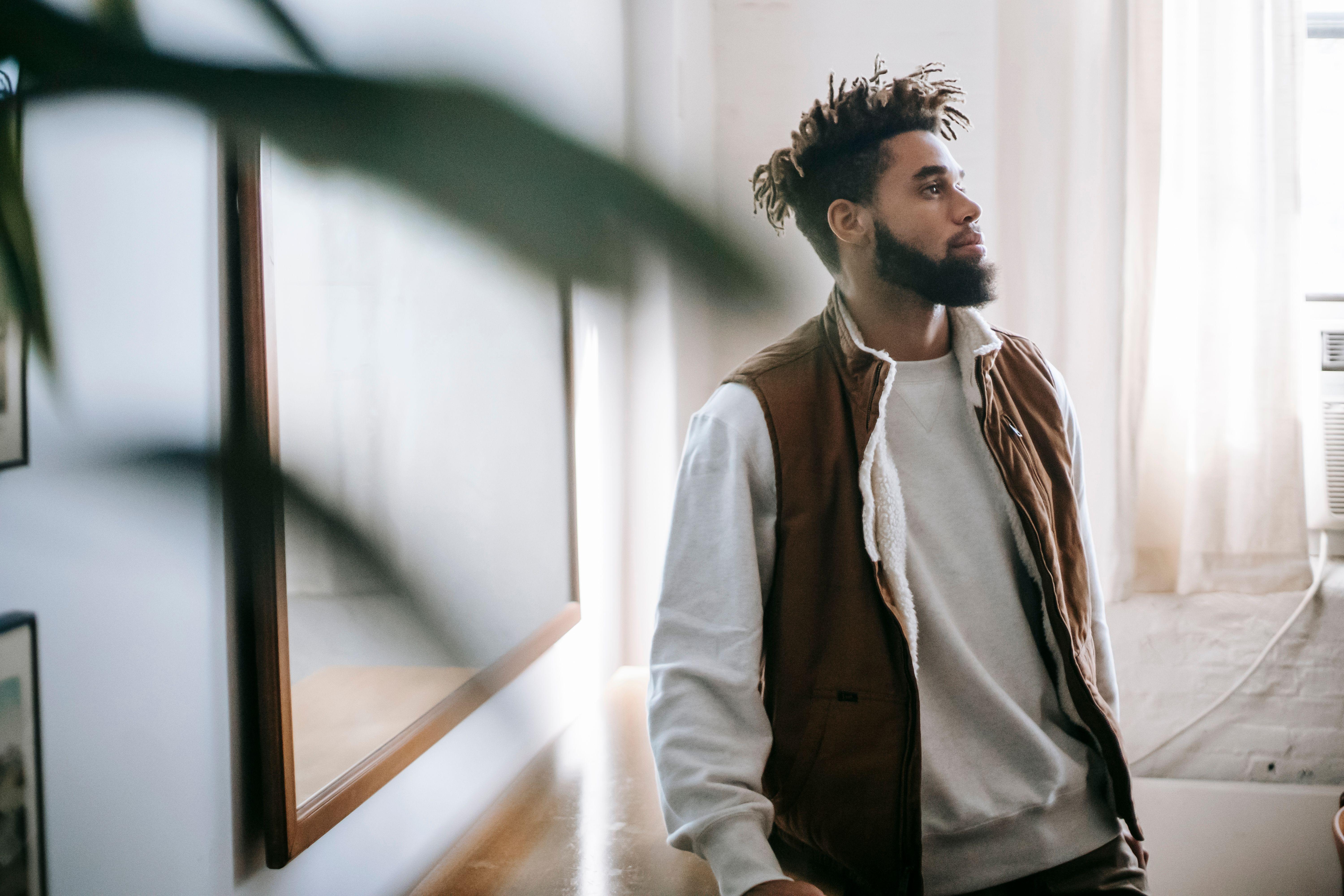 Contemplative young bearded ethnic male millennial in stylish outfit leaning on wooden table near mirror and looking away at home