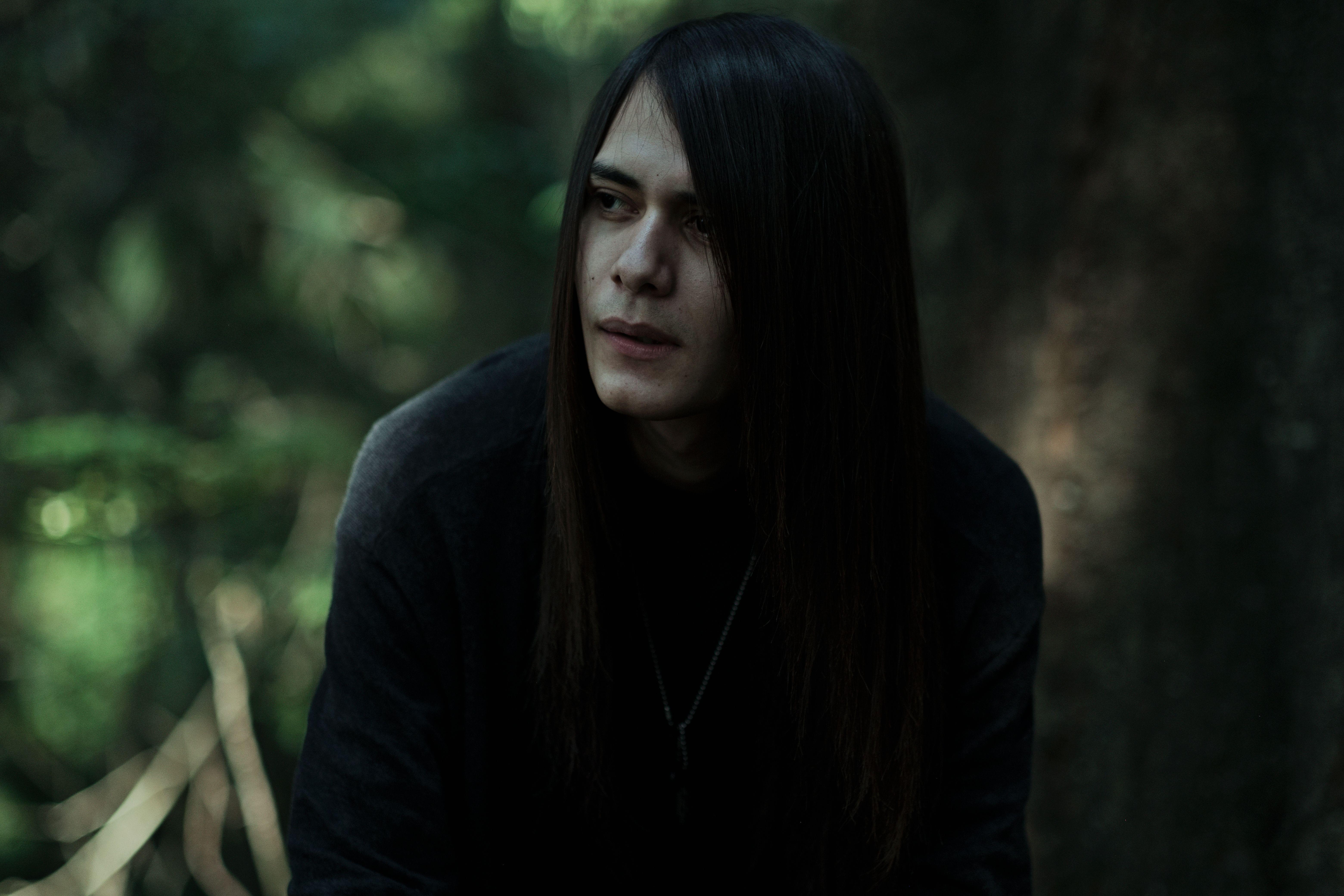 A thoughtful man with long hair poses in a shadowy forest setting.