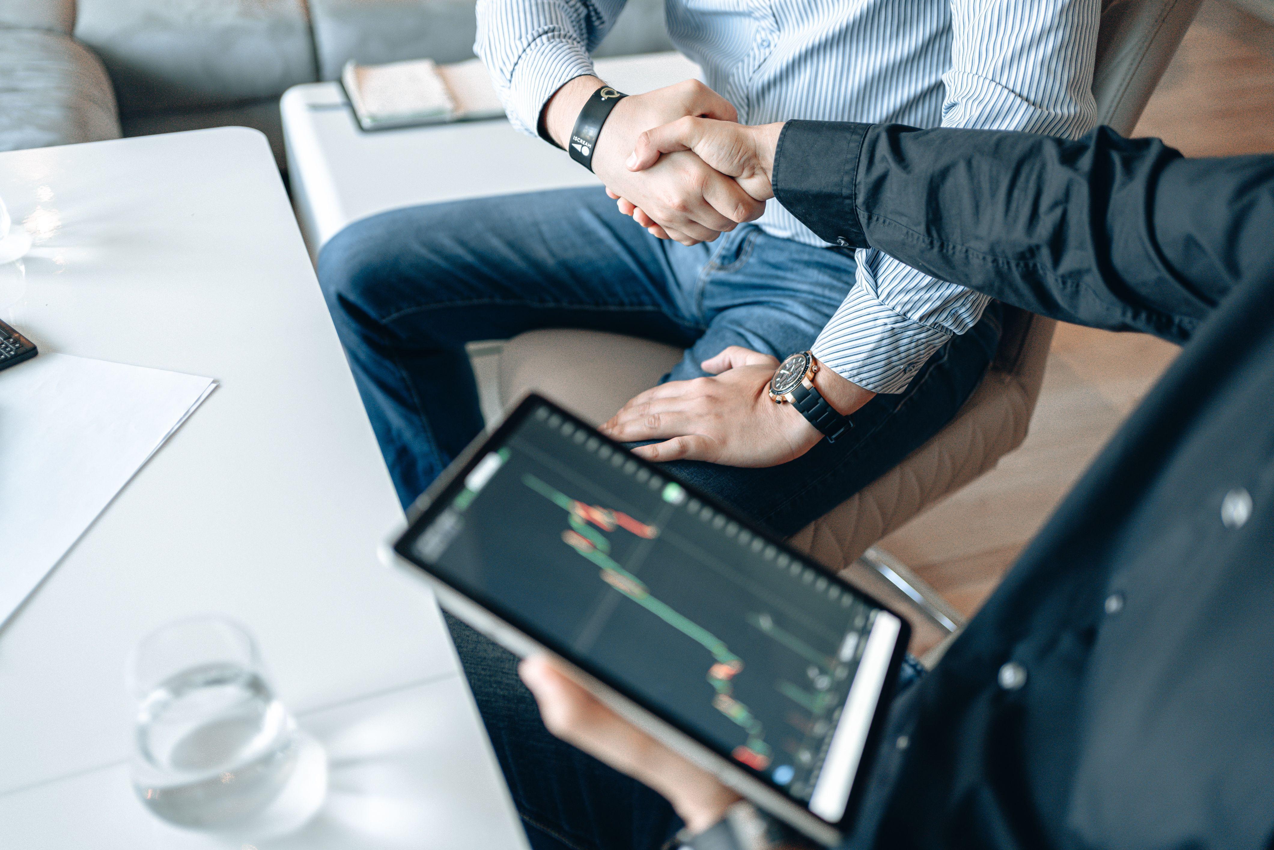 Business professionals shaking hands with a tablet displaying financial graphs.