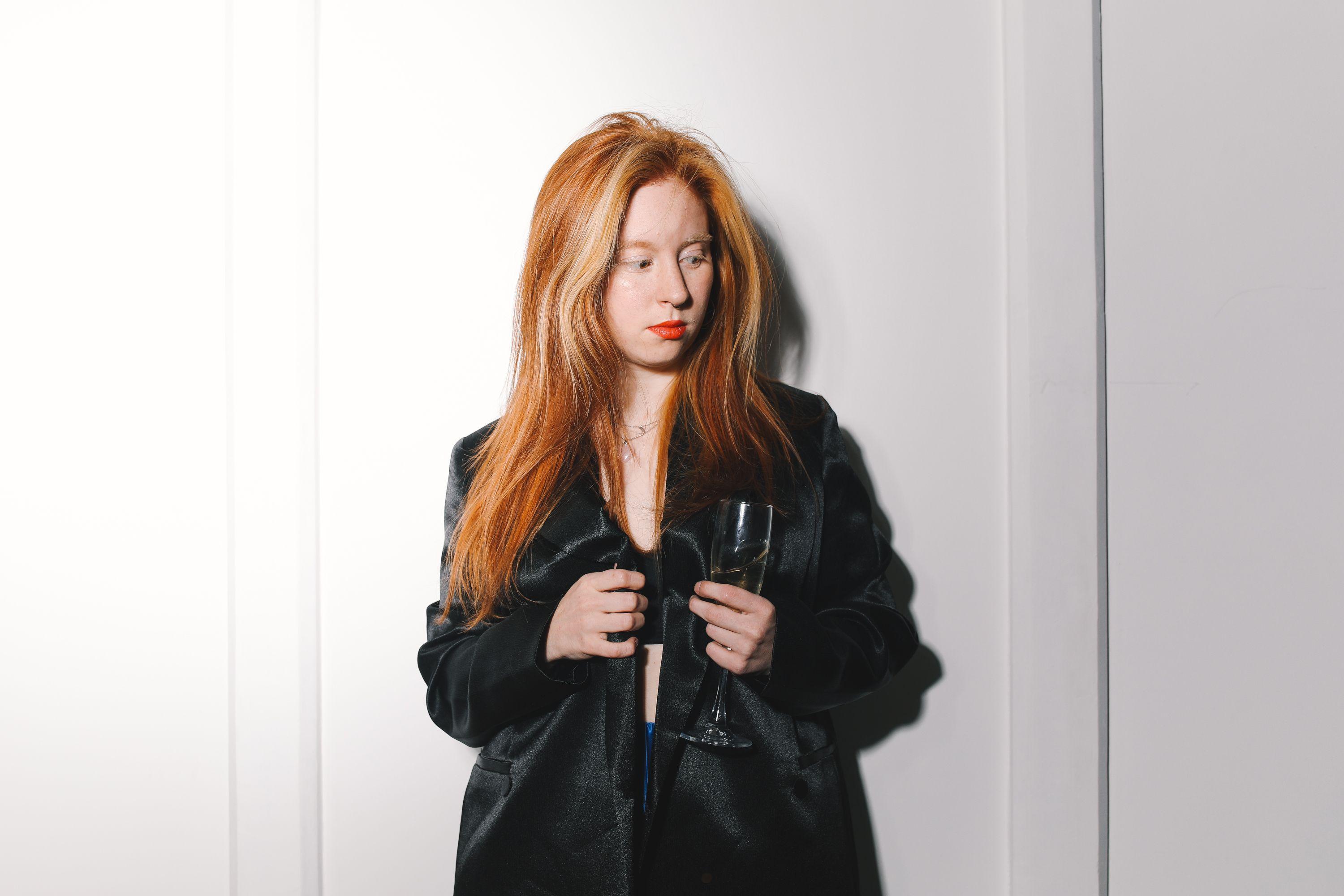 A stylish woman with red hair poses gracefully in a black jacket holding a wine glass.
