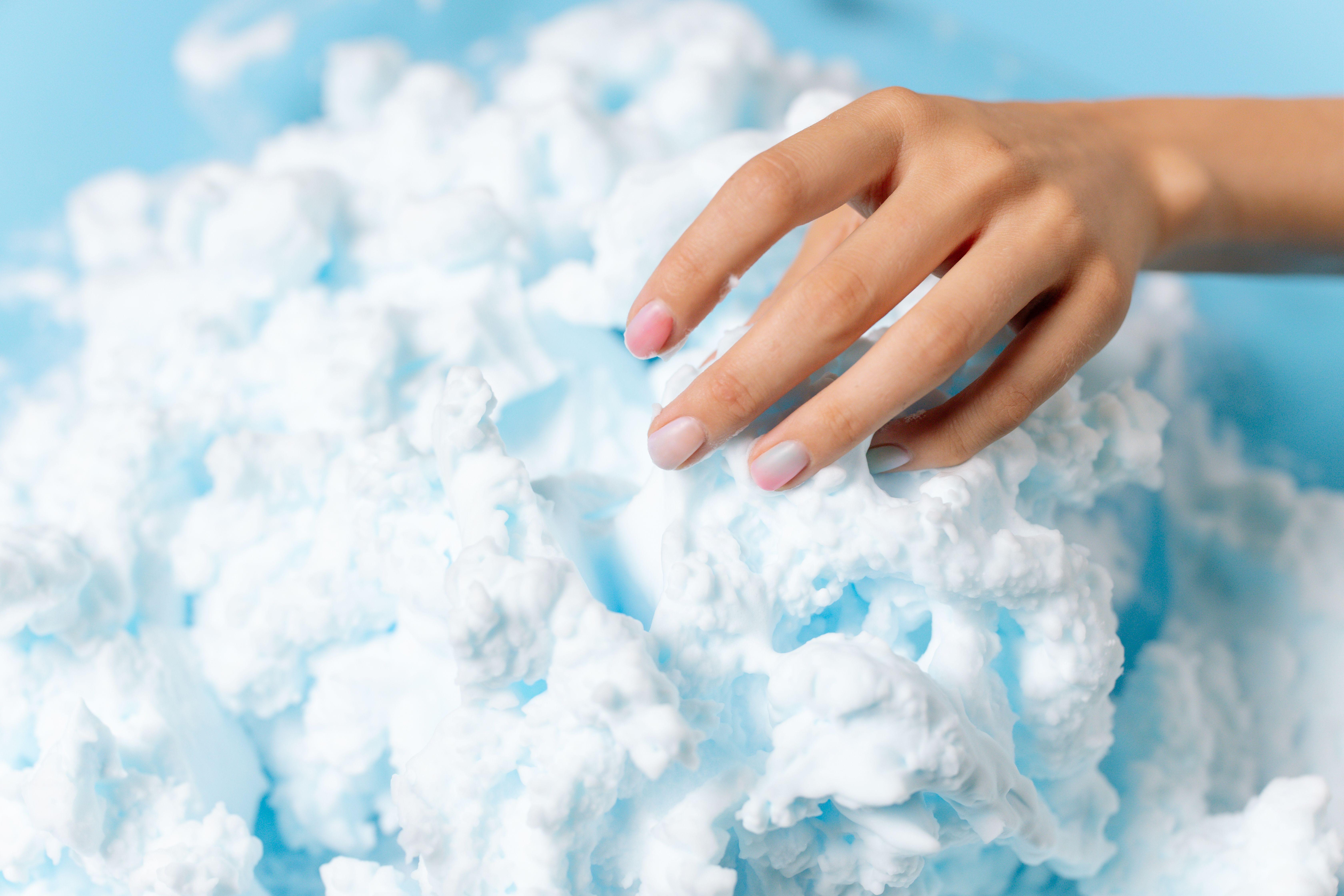 A hand gently touches fluffy blue foam resembling clouds, creating a conceptual abstract image.