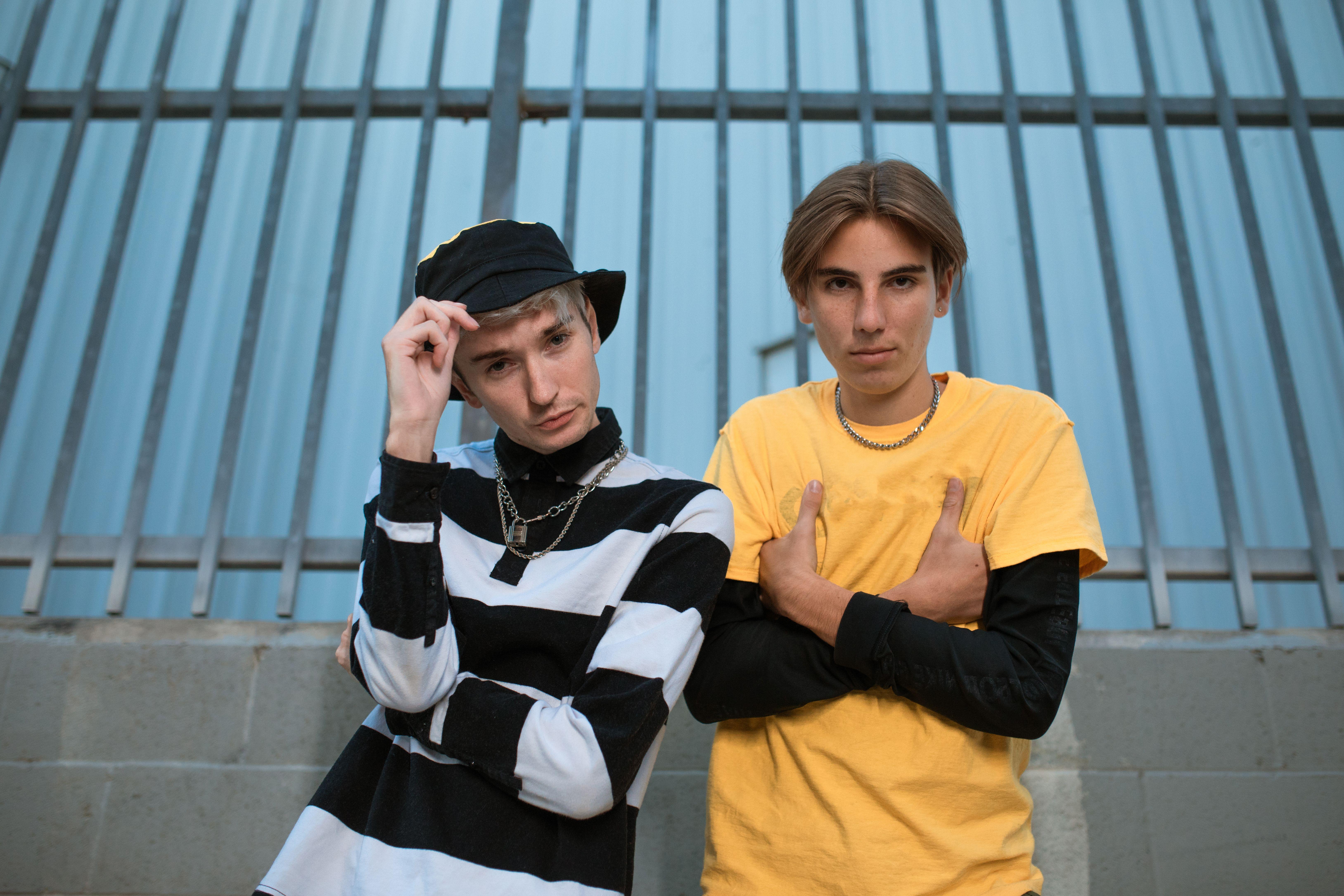 Two trendy teenagers posing in casual outfits against a metal fence background.