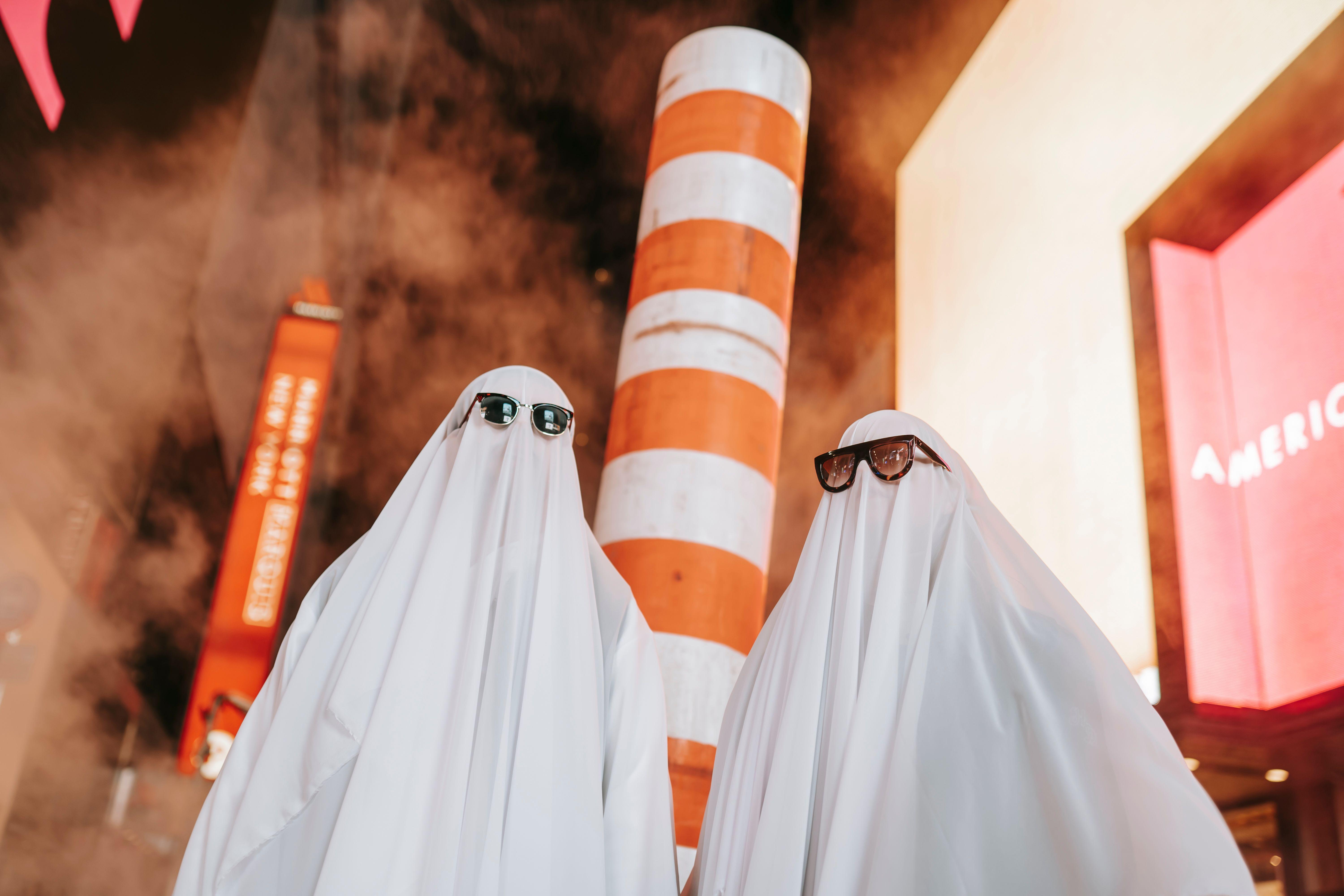From below of anonymous couple wearing ghost costumes and sunglasses standing near steam pipe and glowing signboard on building at night time during Halloween holiday