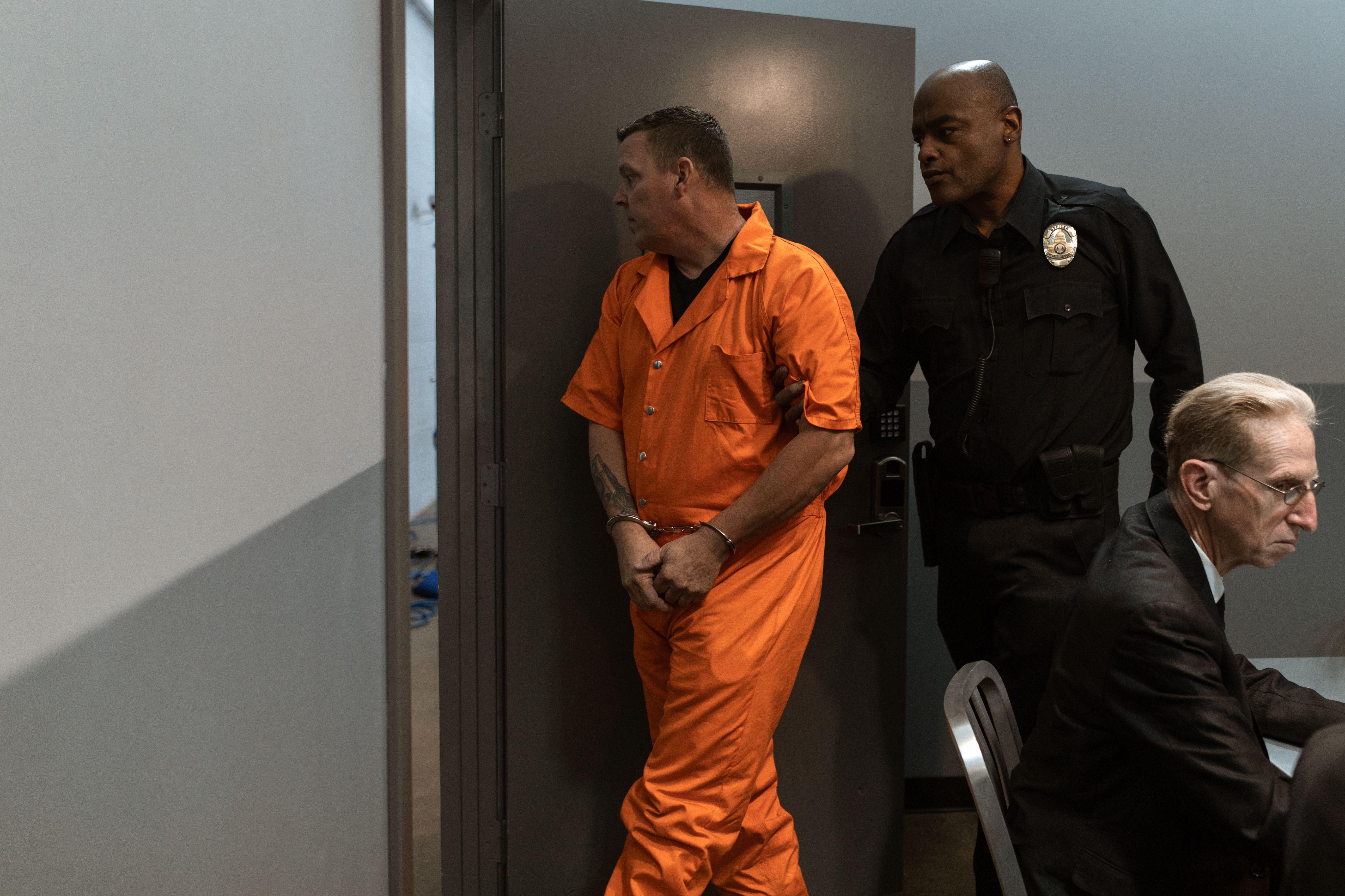 Prisoner in orange jumpsuit being escorted by a correctional officer through facility.