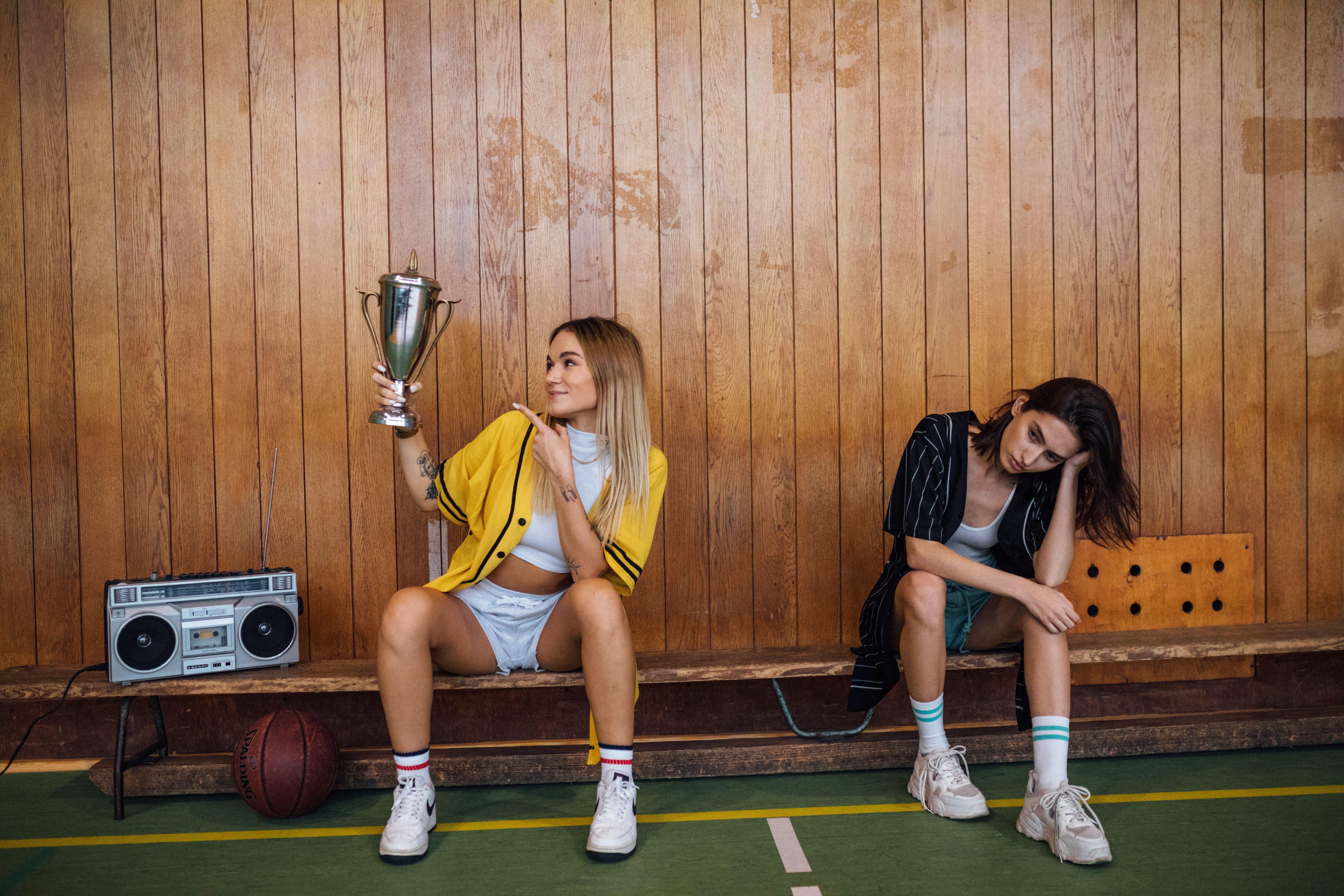 Two young women on a bench, one celebrating with a trophy, depicting competition.