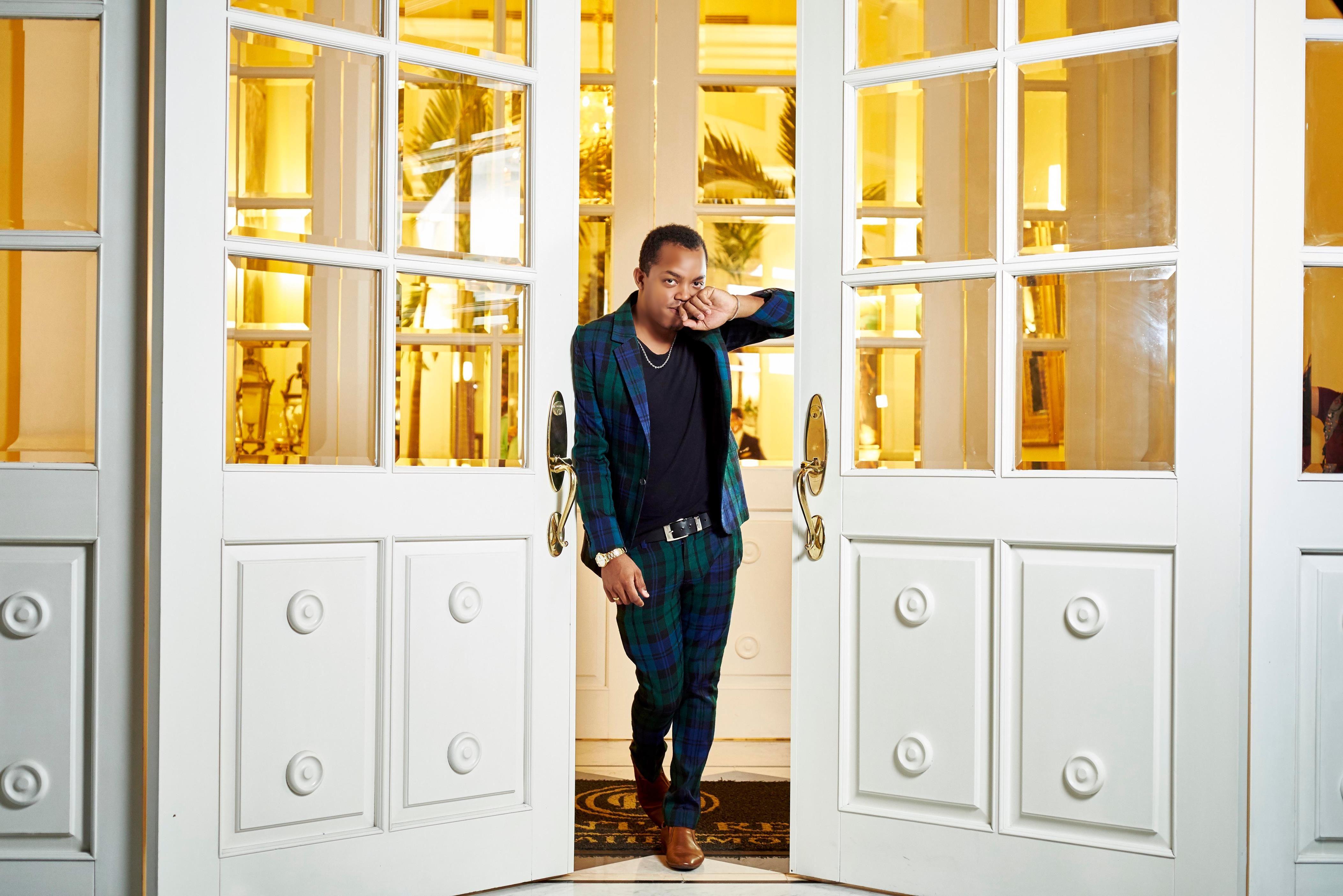 A stylish man in a plaid suit stands confidently at a luxurious entrance with open doors.