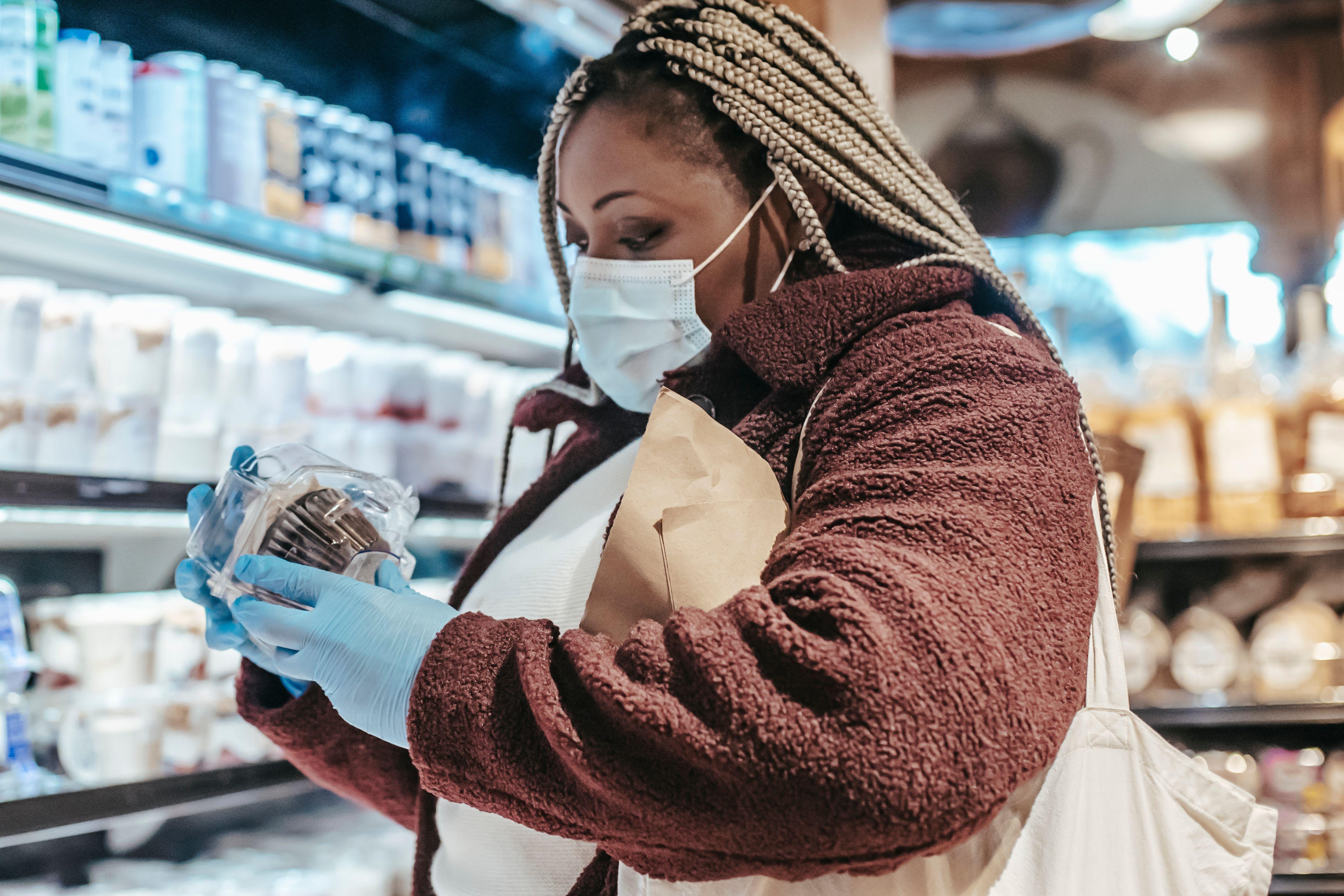 Side view of African American woman in protective gloves and mask reading sticker on product in grocery store