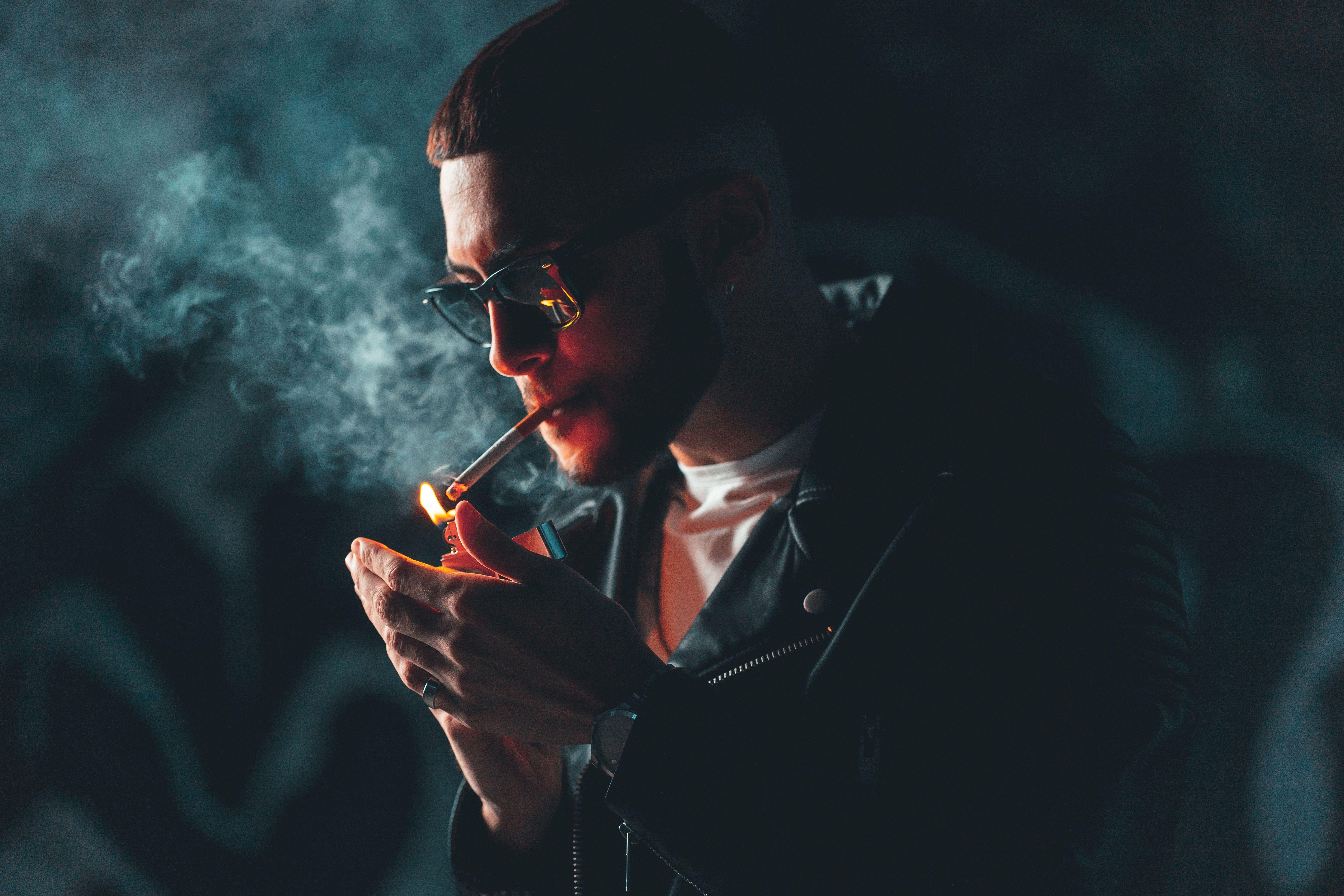 Close-up of a man in sunglasses lighting a cigarette in a dark setting, exuding a moody vibe.