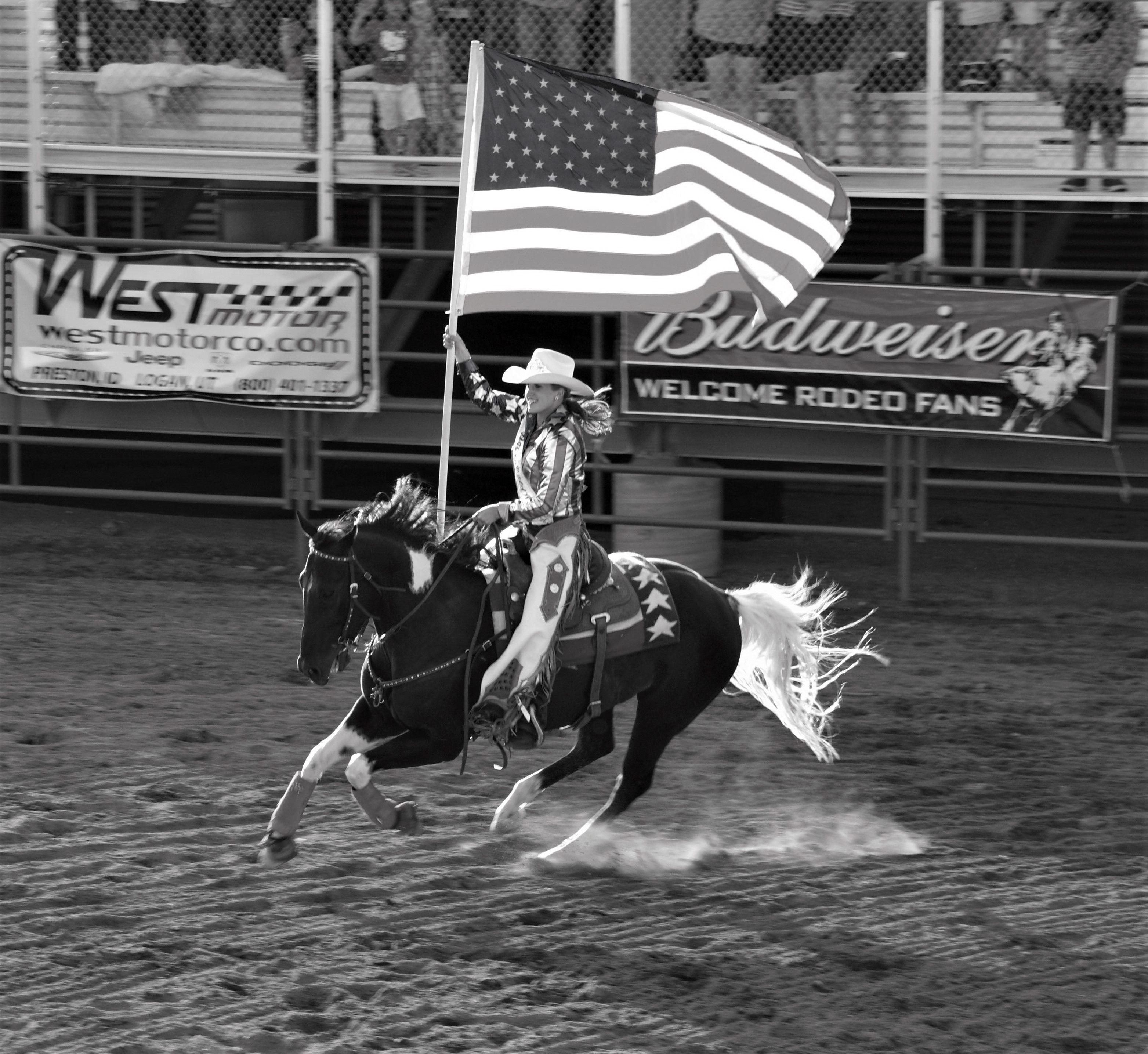 A cowgirl rides a horse with the American flag at a rodeo event. Black and white action shot.