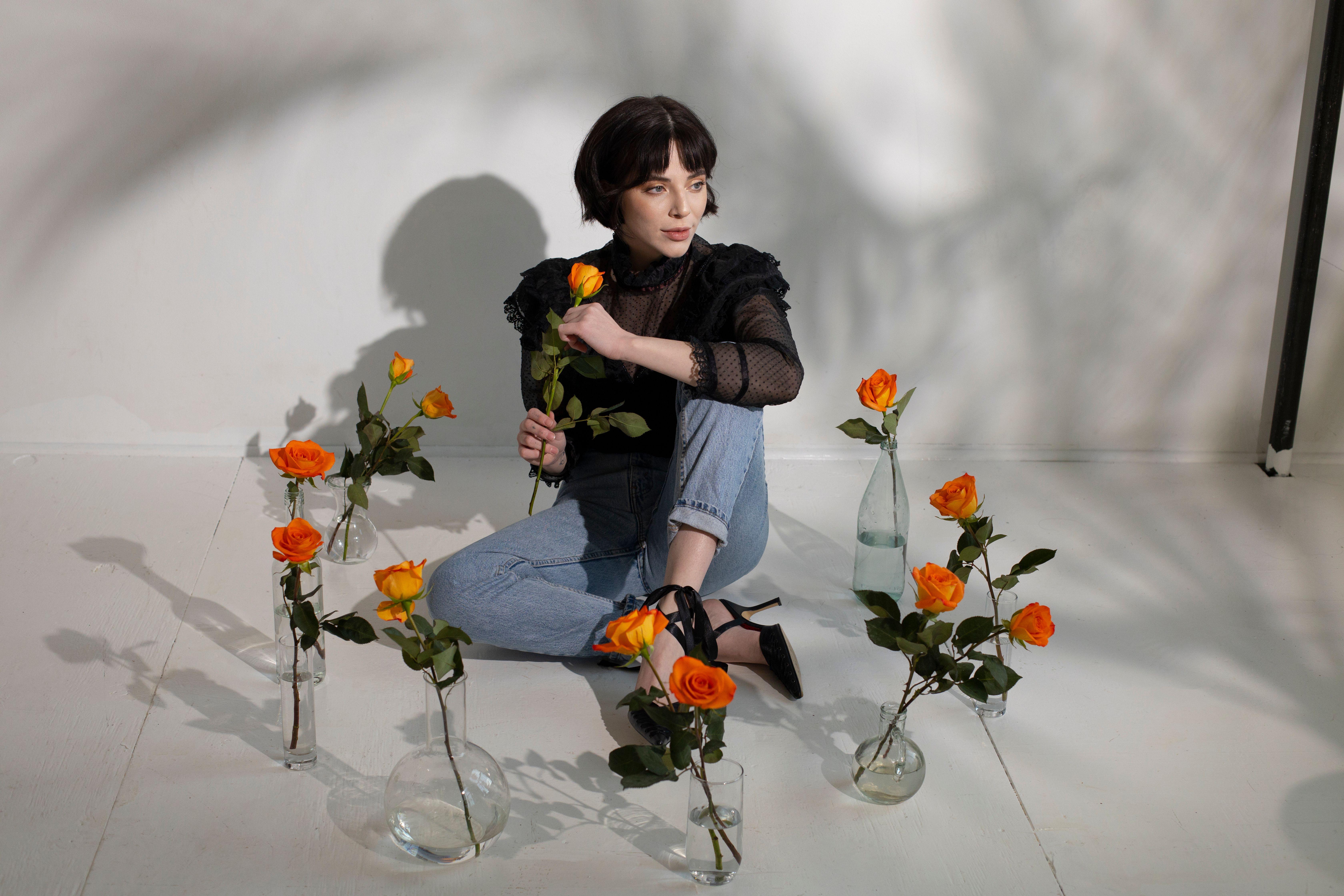 Full length of young dreamy female in stylish outfit sitting on tile floor with glass vases with orange roses in water near white wall with shades while looking away