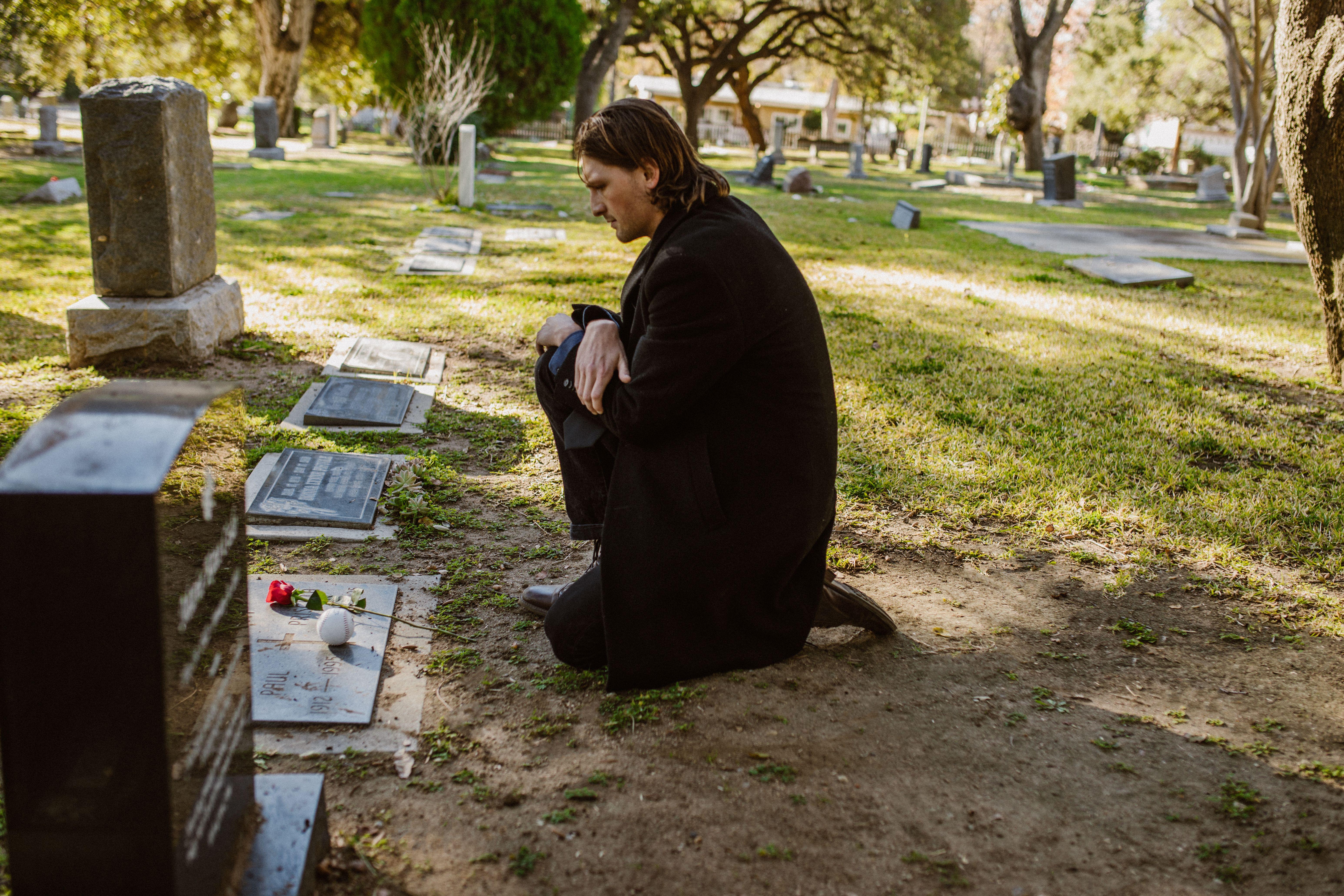 A man kneeling at a gravestone in a cemetery, holding a red rose during an outdoor visit.