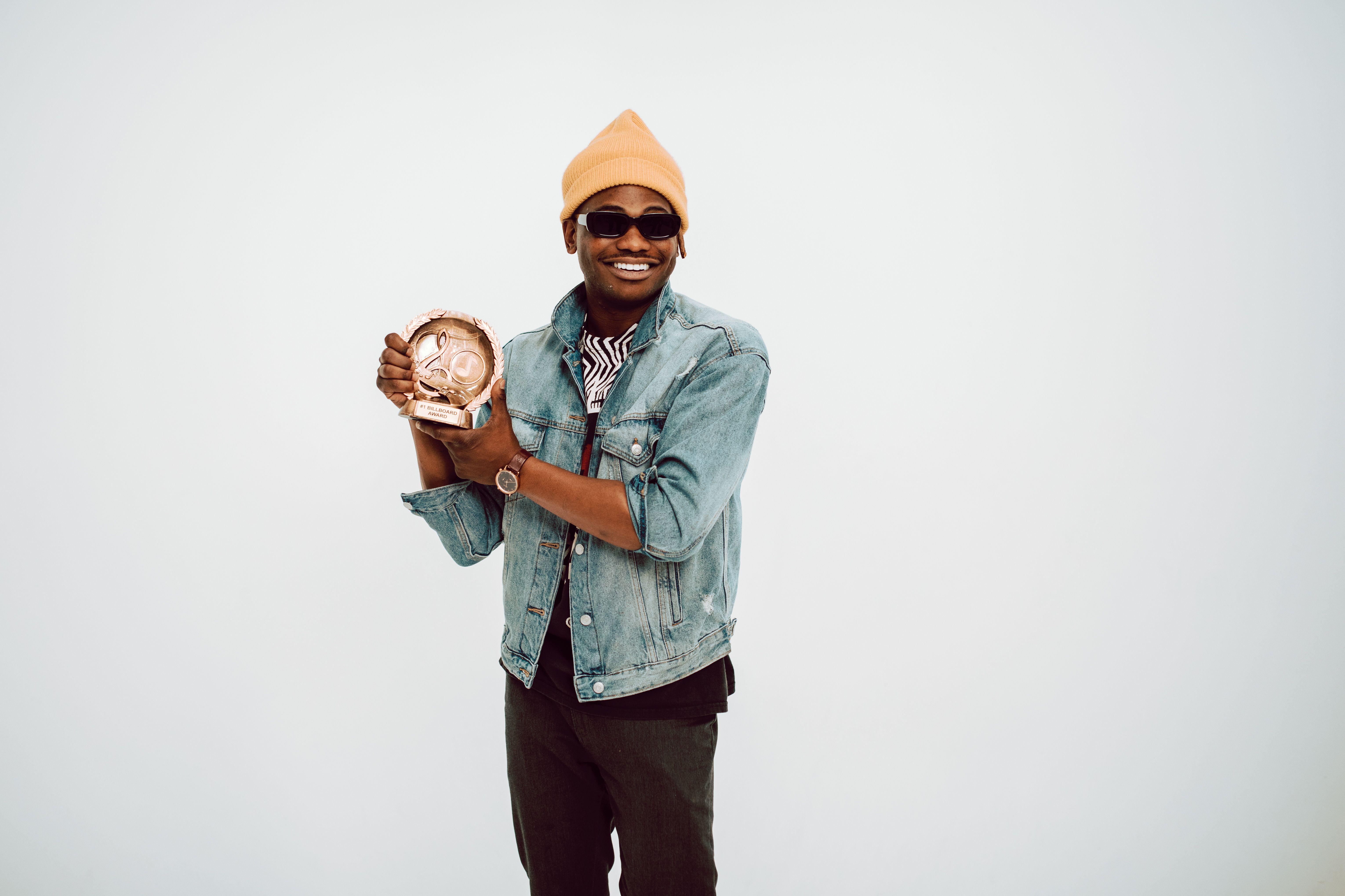 Smiling man in casual attire with sunglasses holding a trophy in a studio environment.