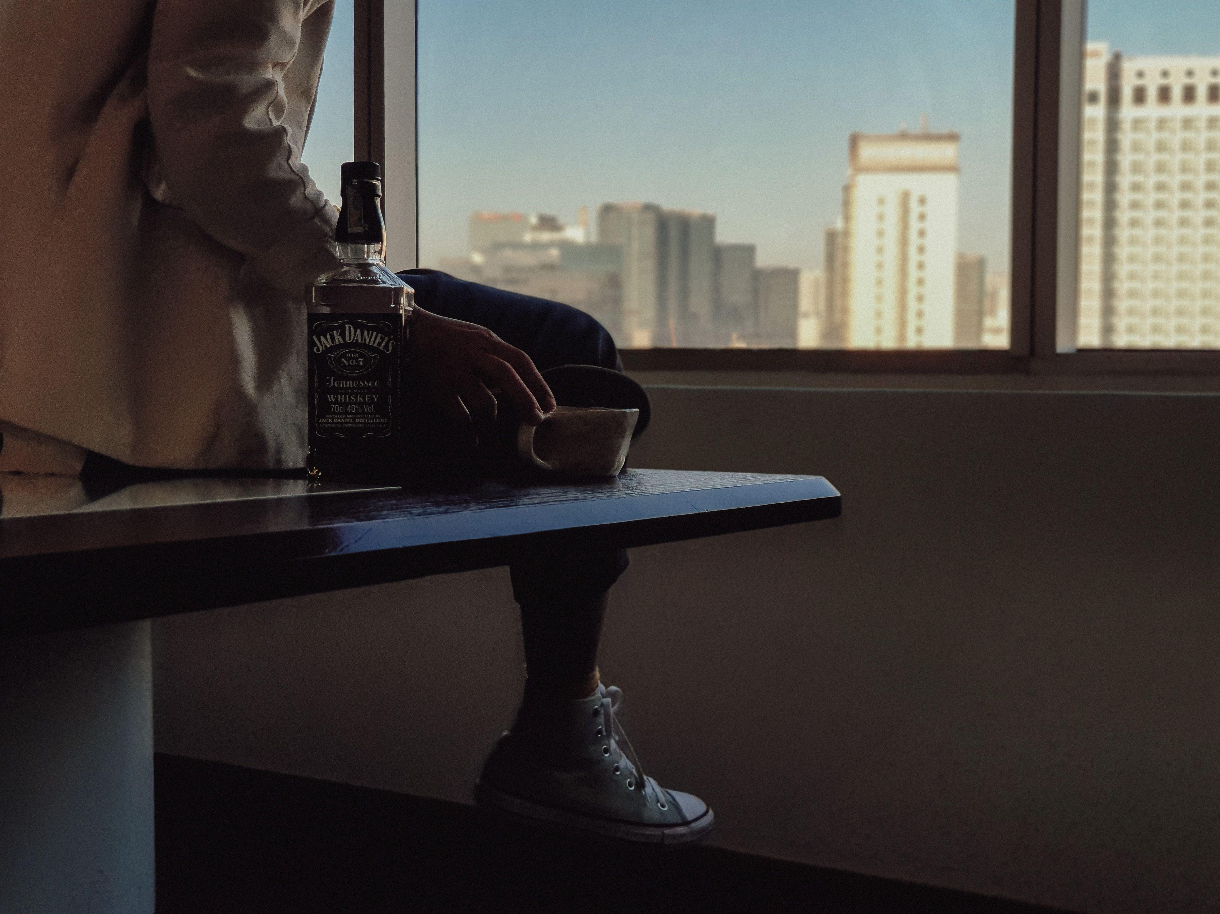 Adult sitting indoors holding a cup beside a bottle of Jack Daniel's, with urban skyline view.
