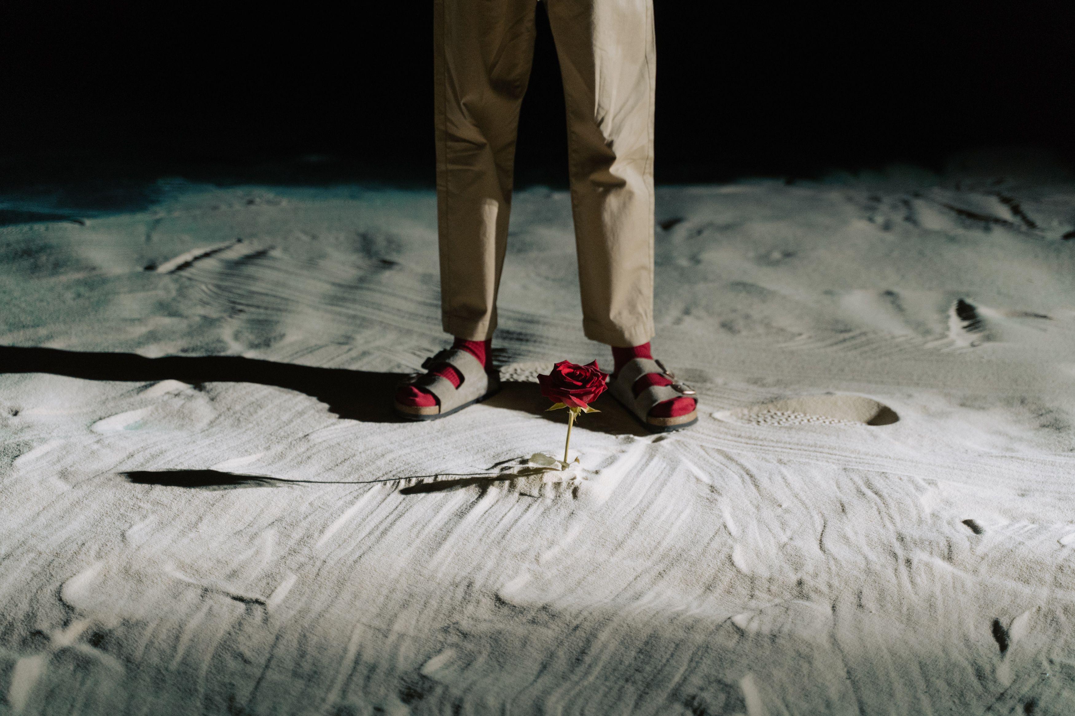 A red rose stands near a person's feet on sandy ground, highlighting contrast and mystery.