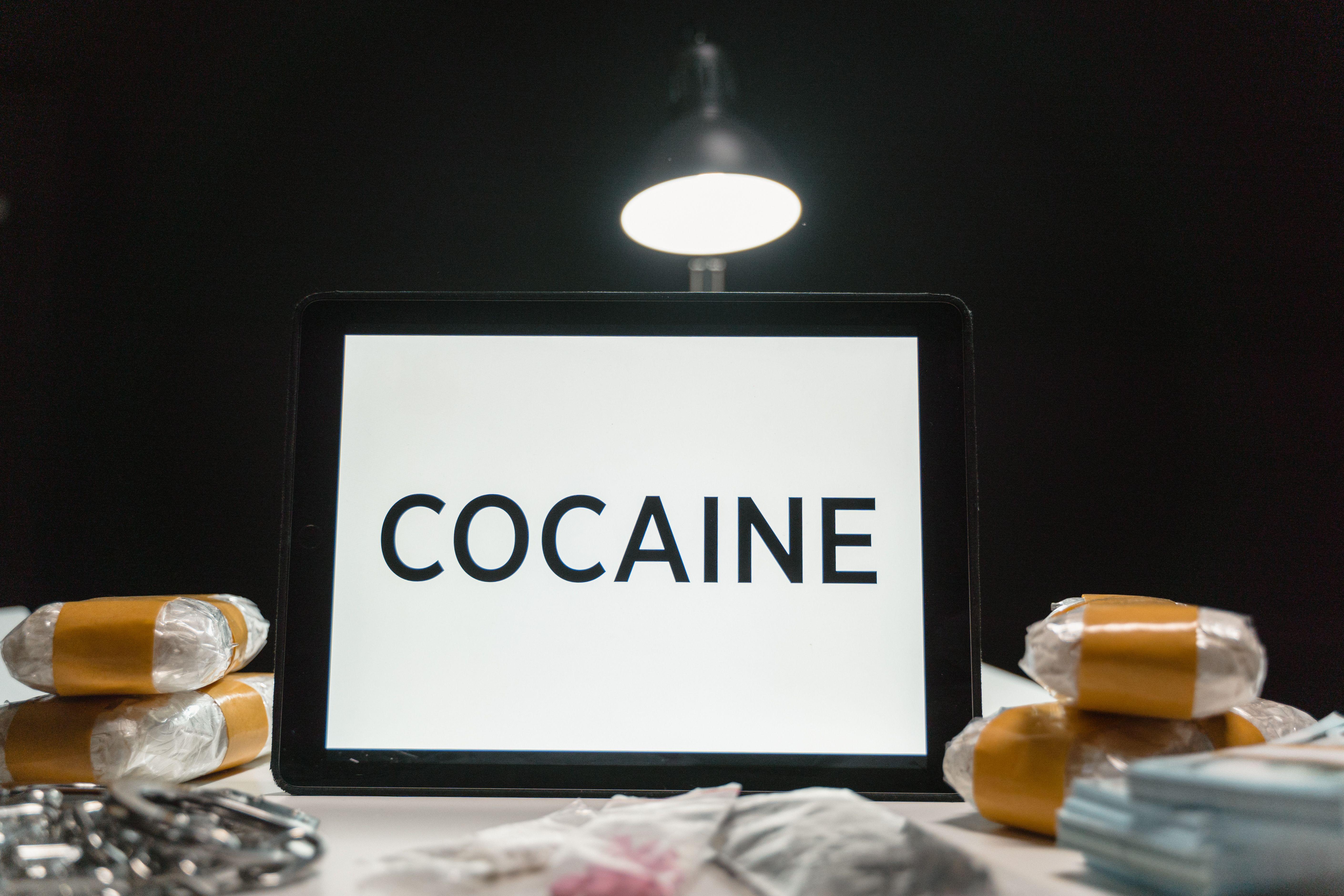Tablet computer displaying the word 'Cocaine' surrounded by wrapped packets in a dimly lit setting.