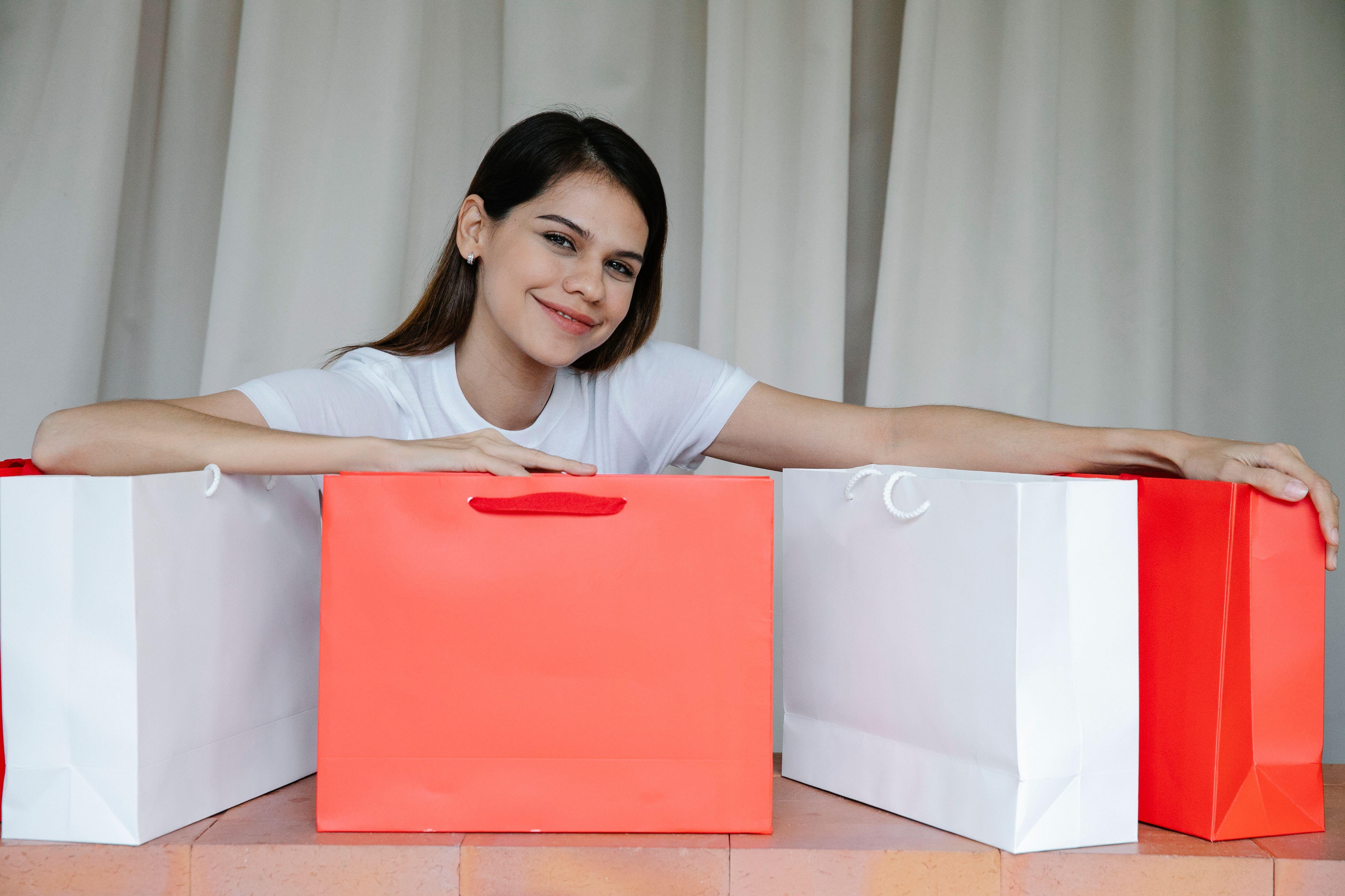 Joyful young lady with long dark hair in white t shirt demonstrating similar paper bags and looking at camera after successful shopping