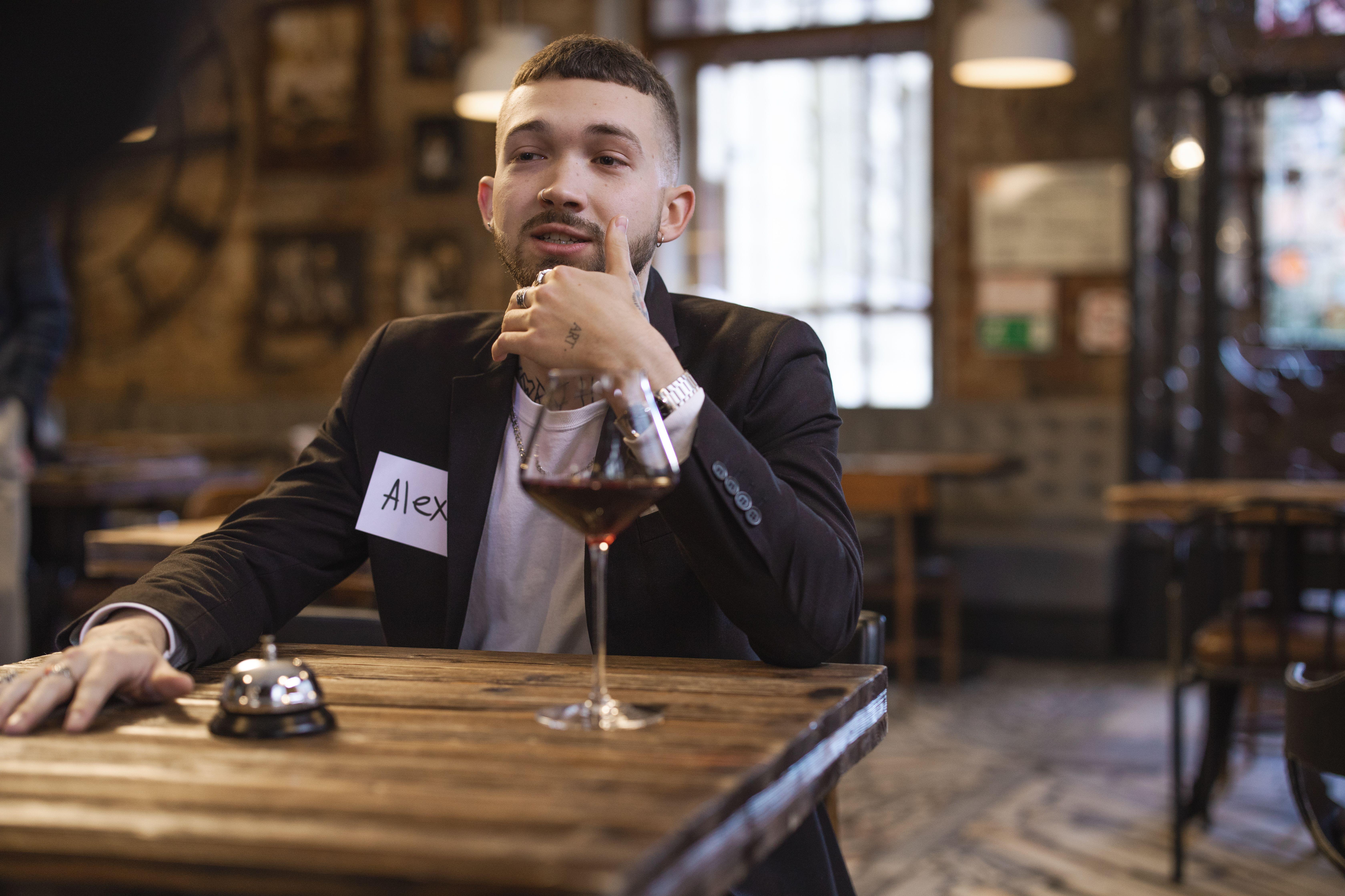 A man with a name tag sits at a table in a rustic bar environment holding a drink.