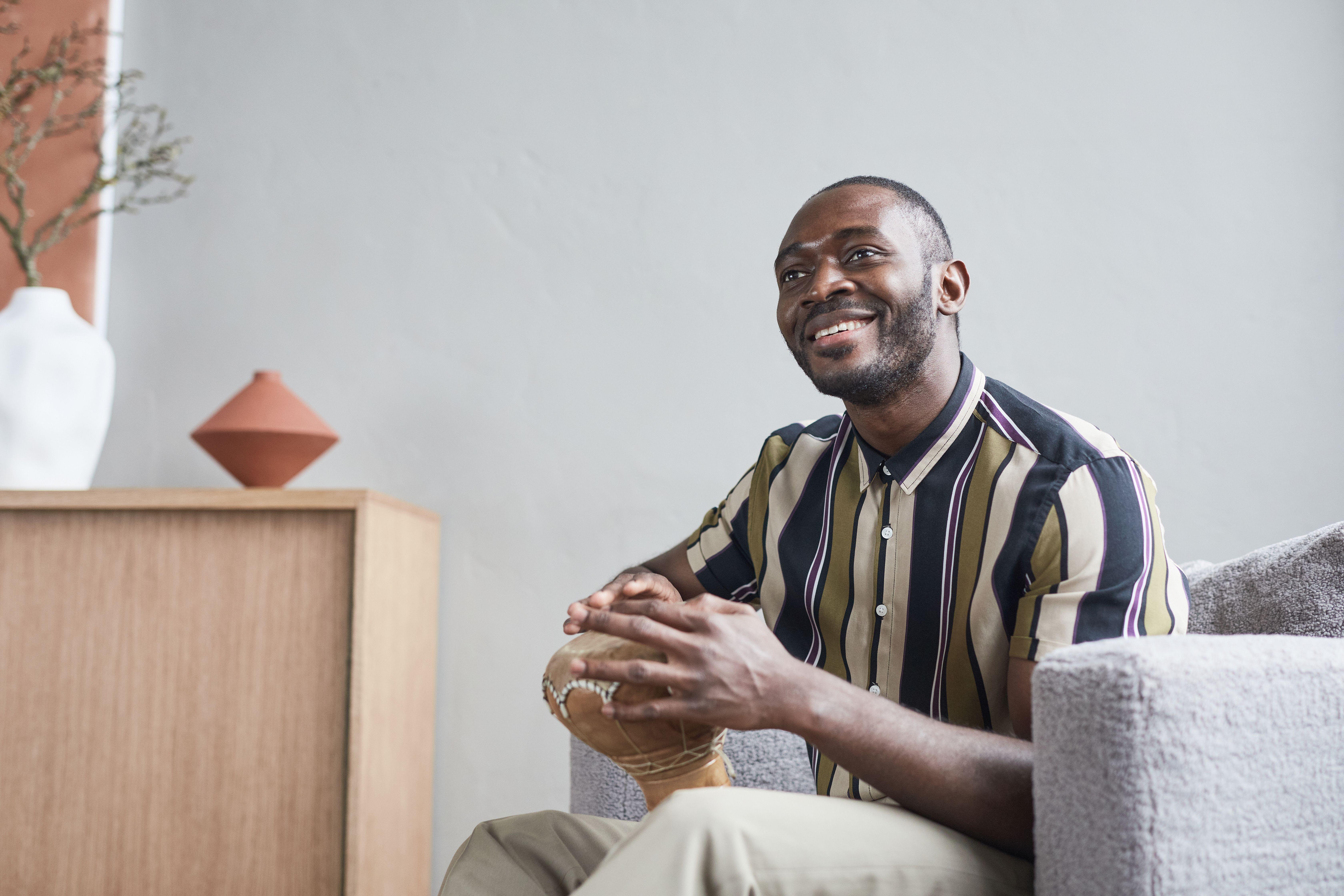 A cheerful African man playing a djembe drum indoors, seated comfortably and smiling.