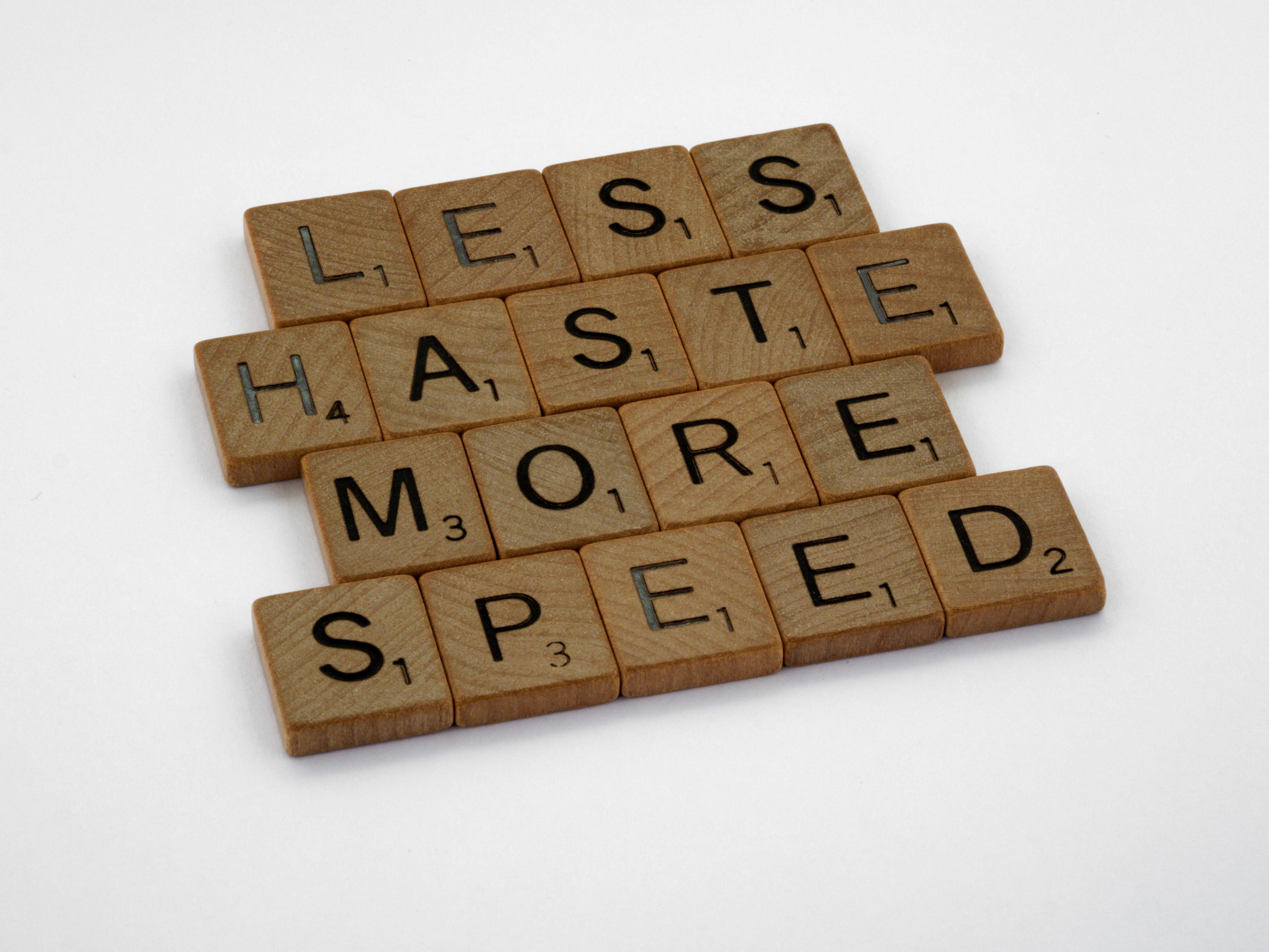 Wooden scrabble tiles forming the phrase 'Less Haste More Speed' on a white background.