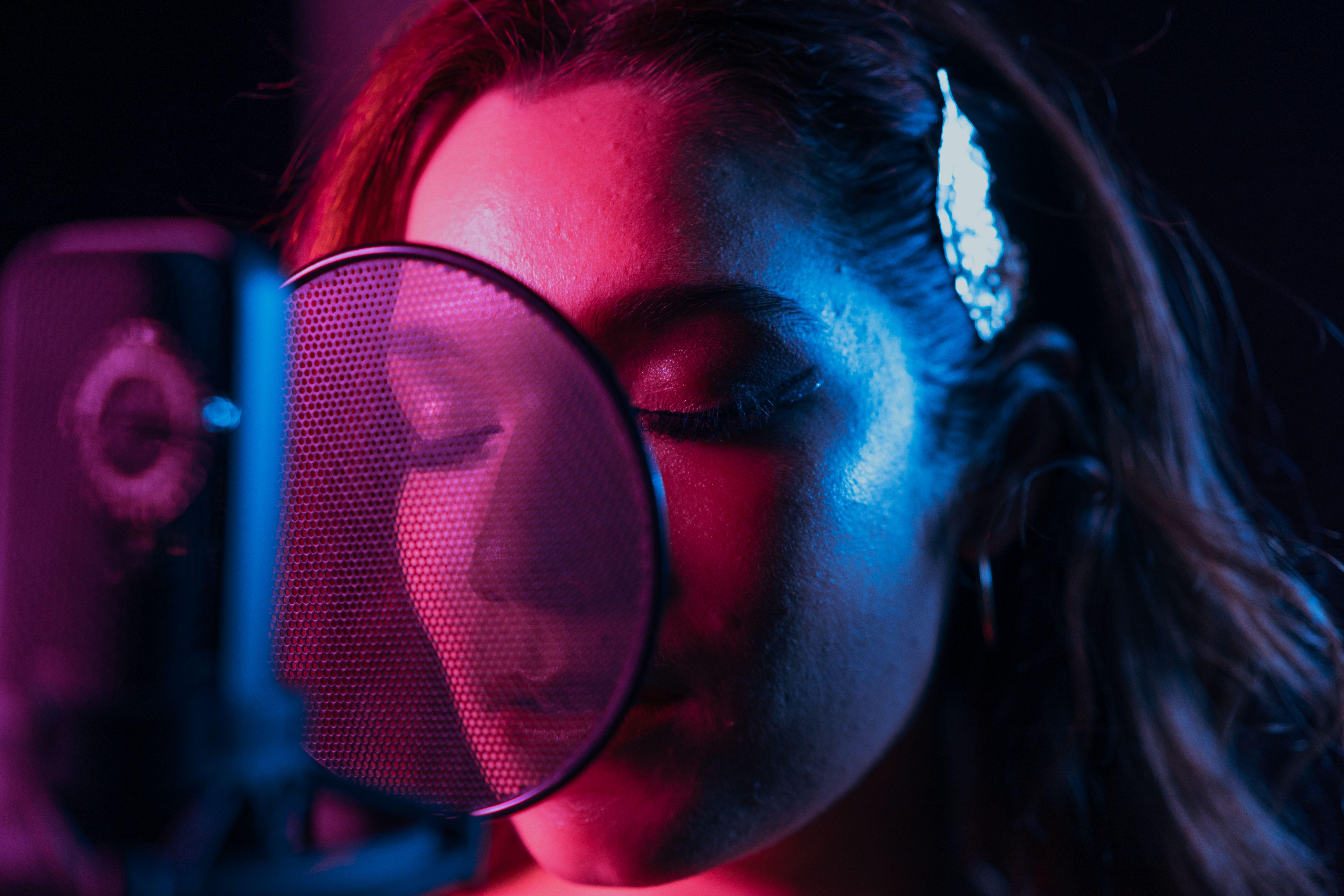 Close-up of a woman singing with eyes closed in a vibrant lit recording studio.