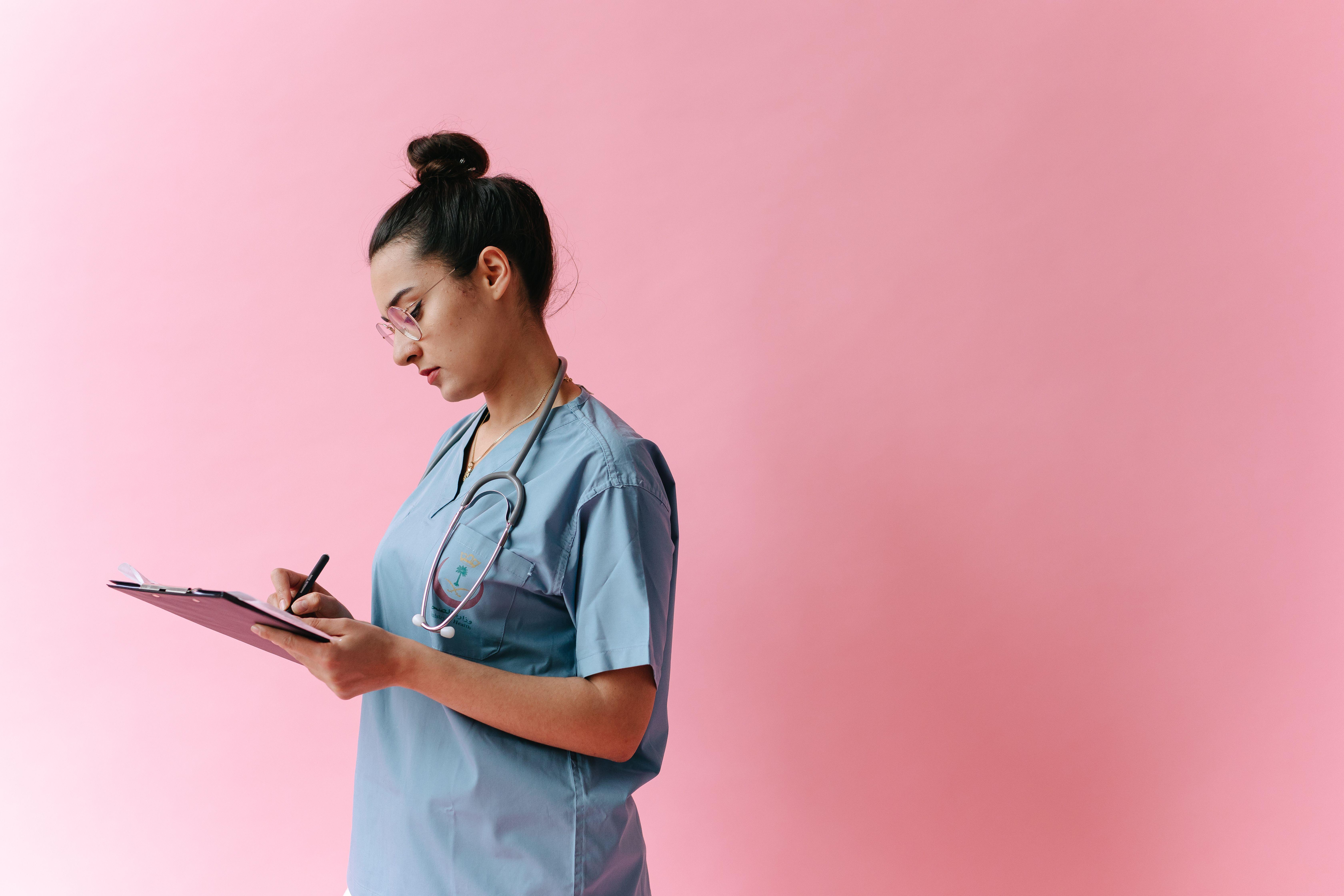 Female nurse in scrubs writing on a clipboard with a stethoscope on a pink background.