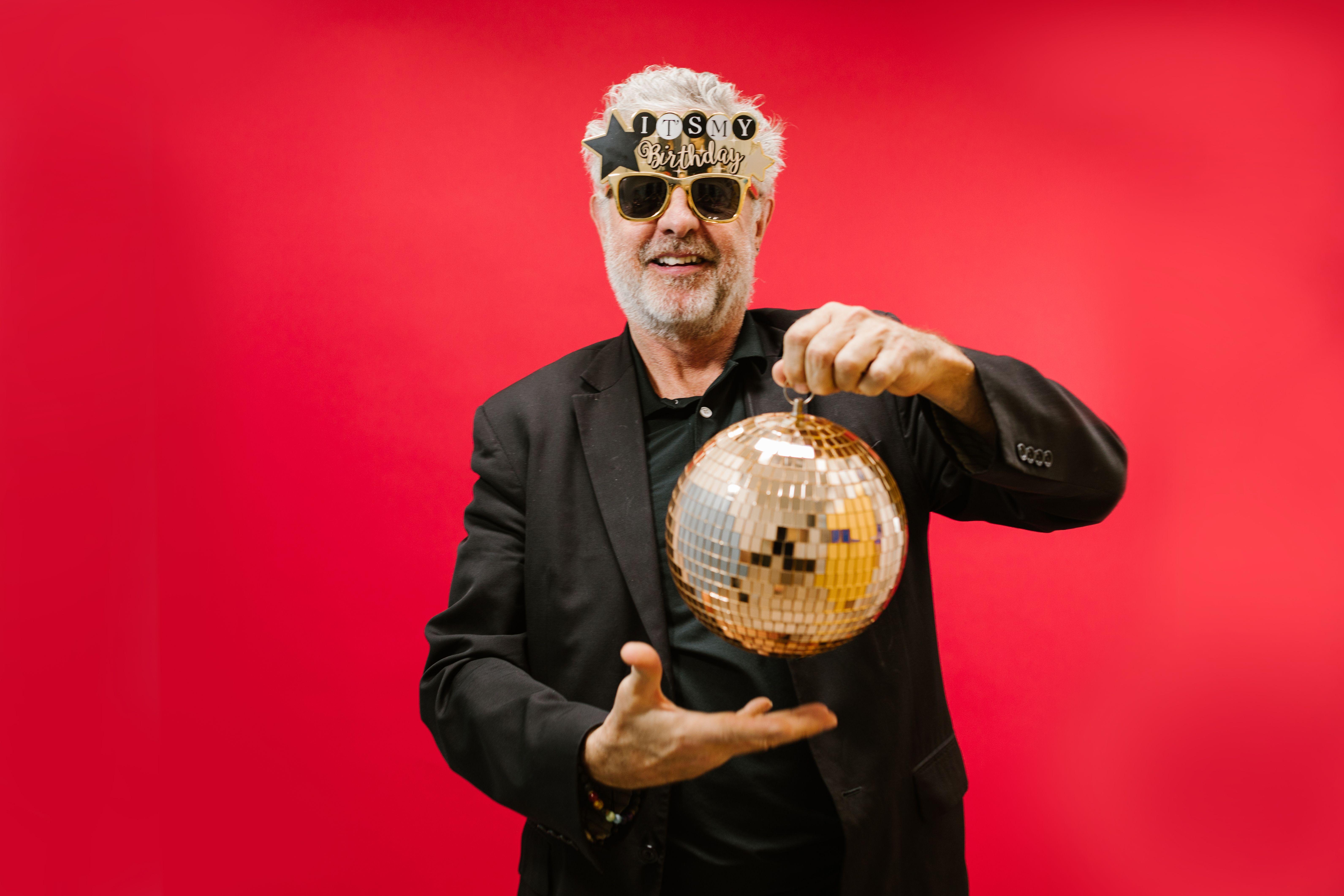 A cheerful elderly man with sunglasses and a disco ball against a vivid red background.