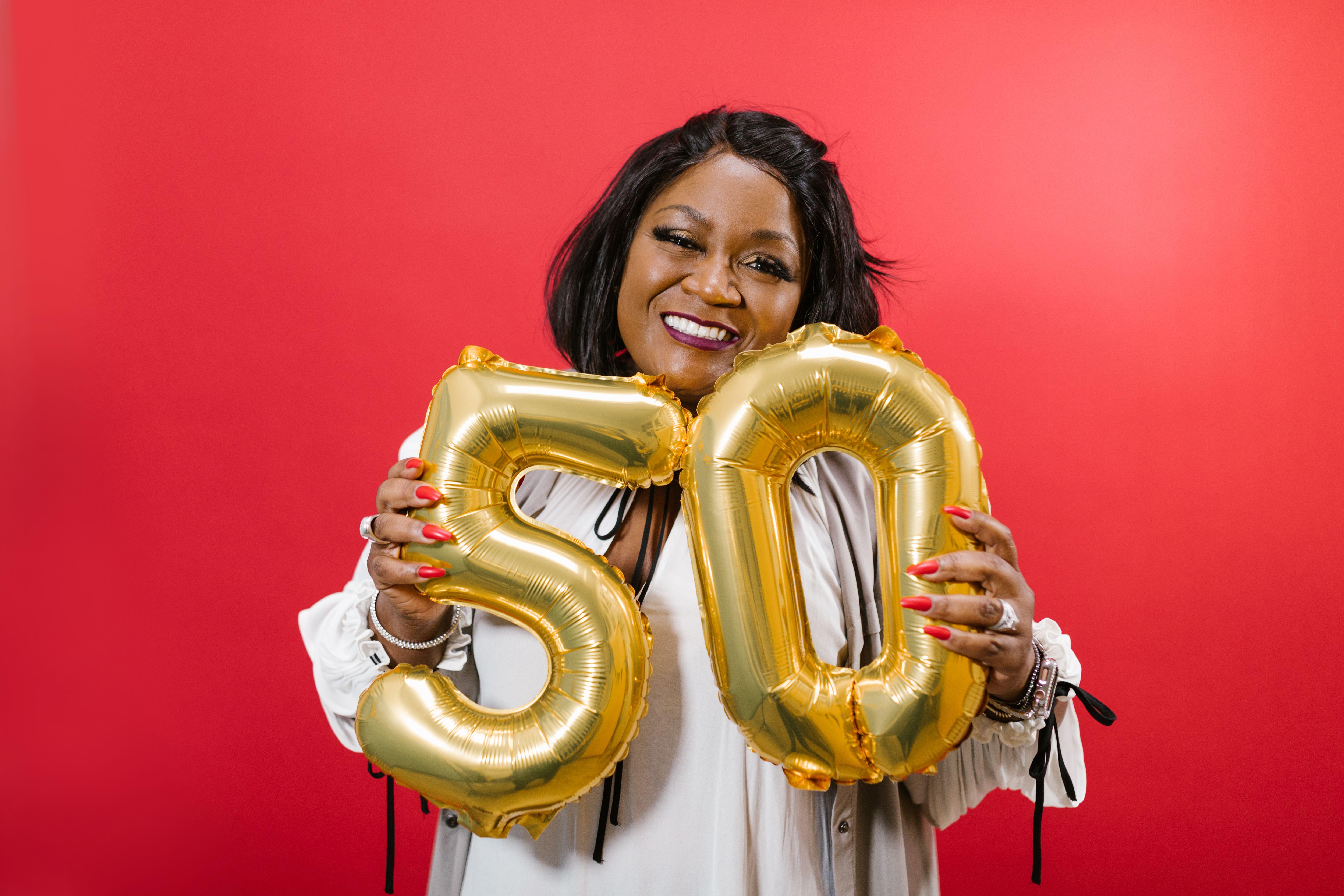 Smiling woman holding golden number 50 balloons against red background.