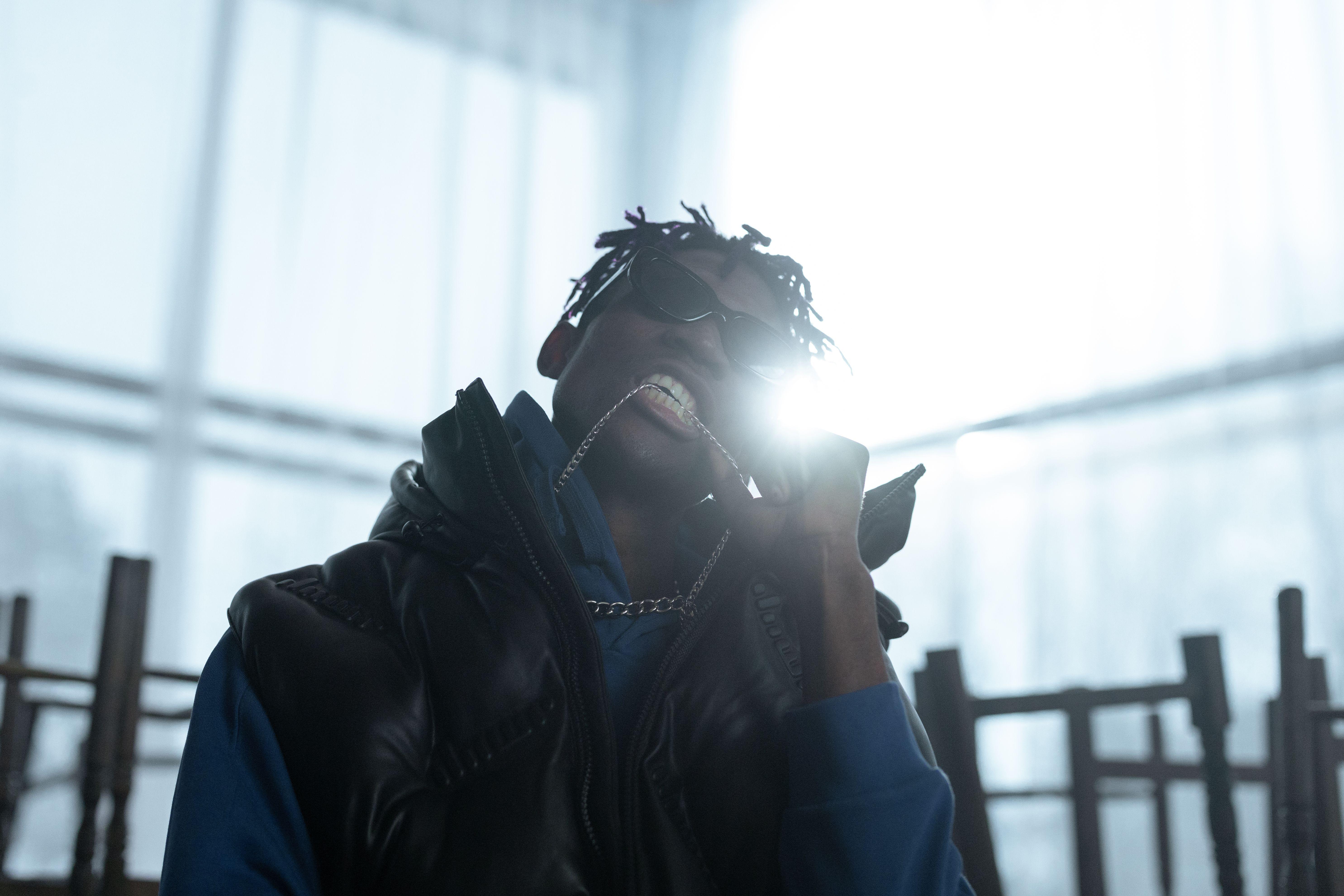 Stylish urban man in sunglasses and streetwear holding a chain indoors with bright backlight.