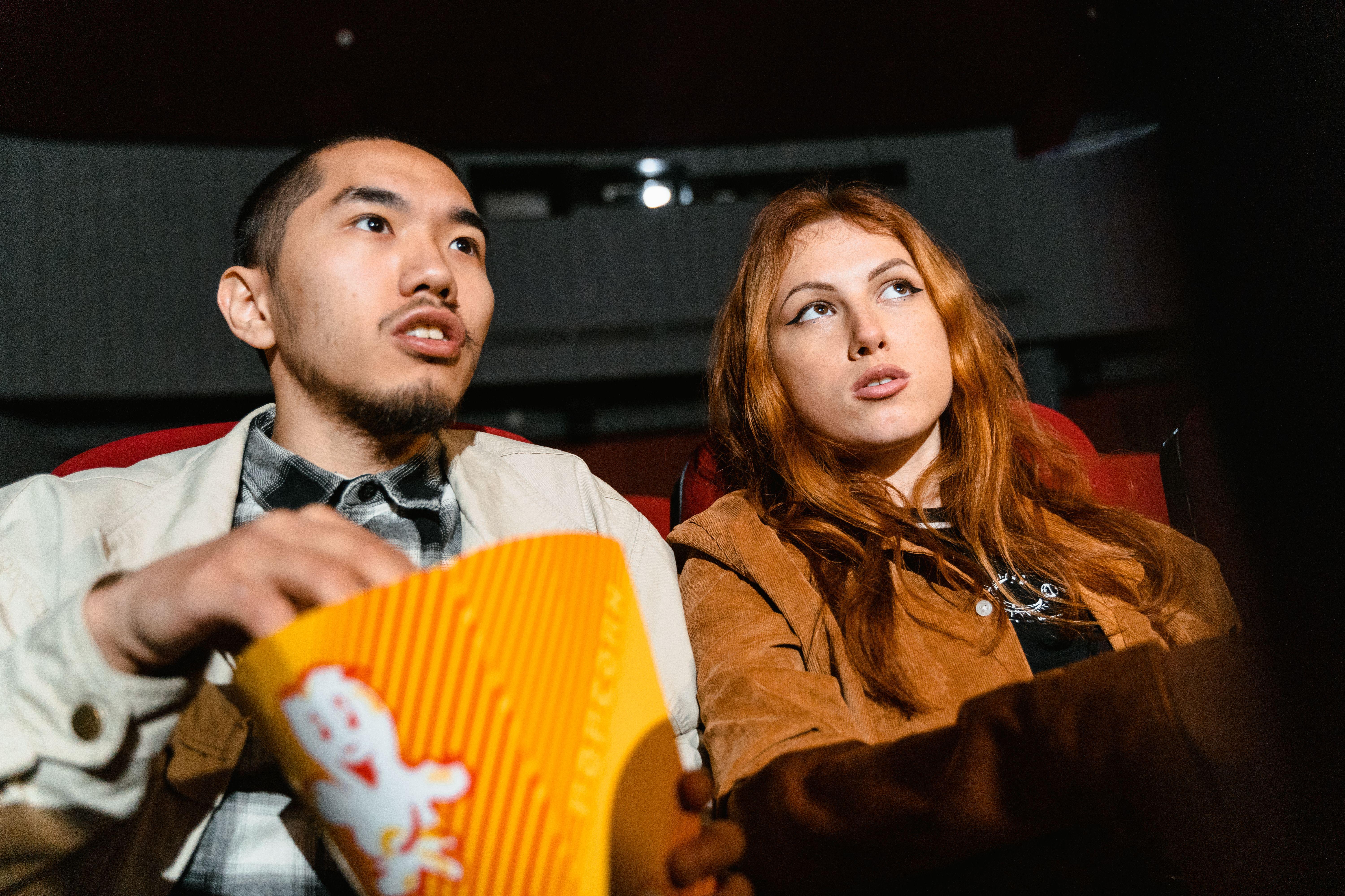 A couple sits in a movie theater, engrossed in a film while enjoying popcorn.